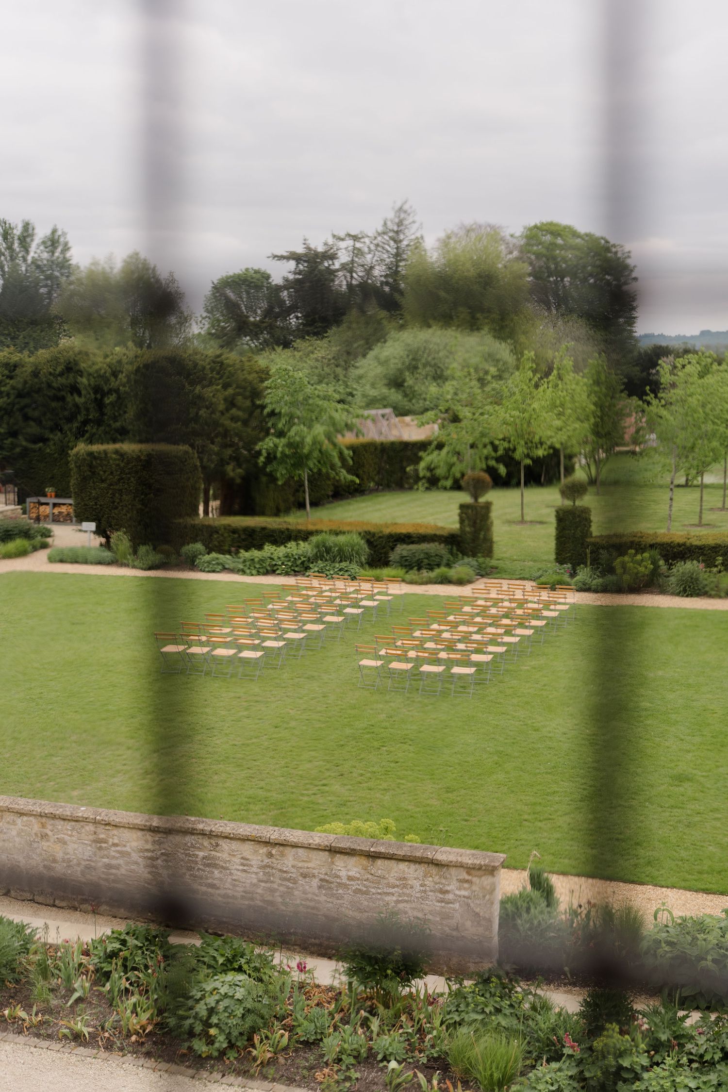 Formal garden with neatly arranged seating on green lawn, surrounded by manicured hedges and trees under overcast sky.