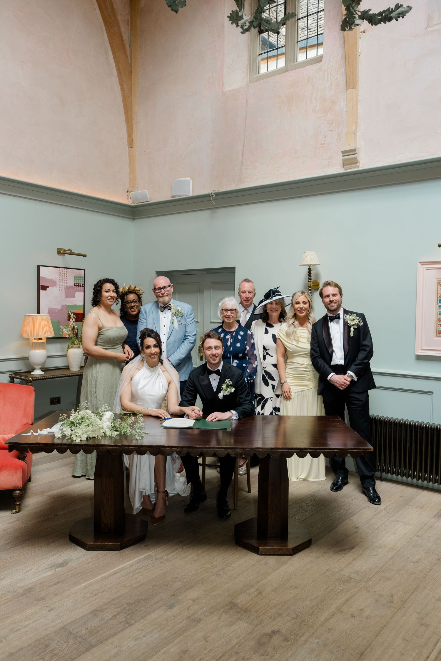 Bride and groom sign wedding register at wooden table surrounded by family and wedding party in elegant venue.