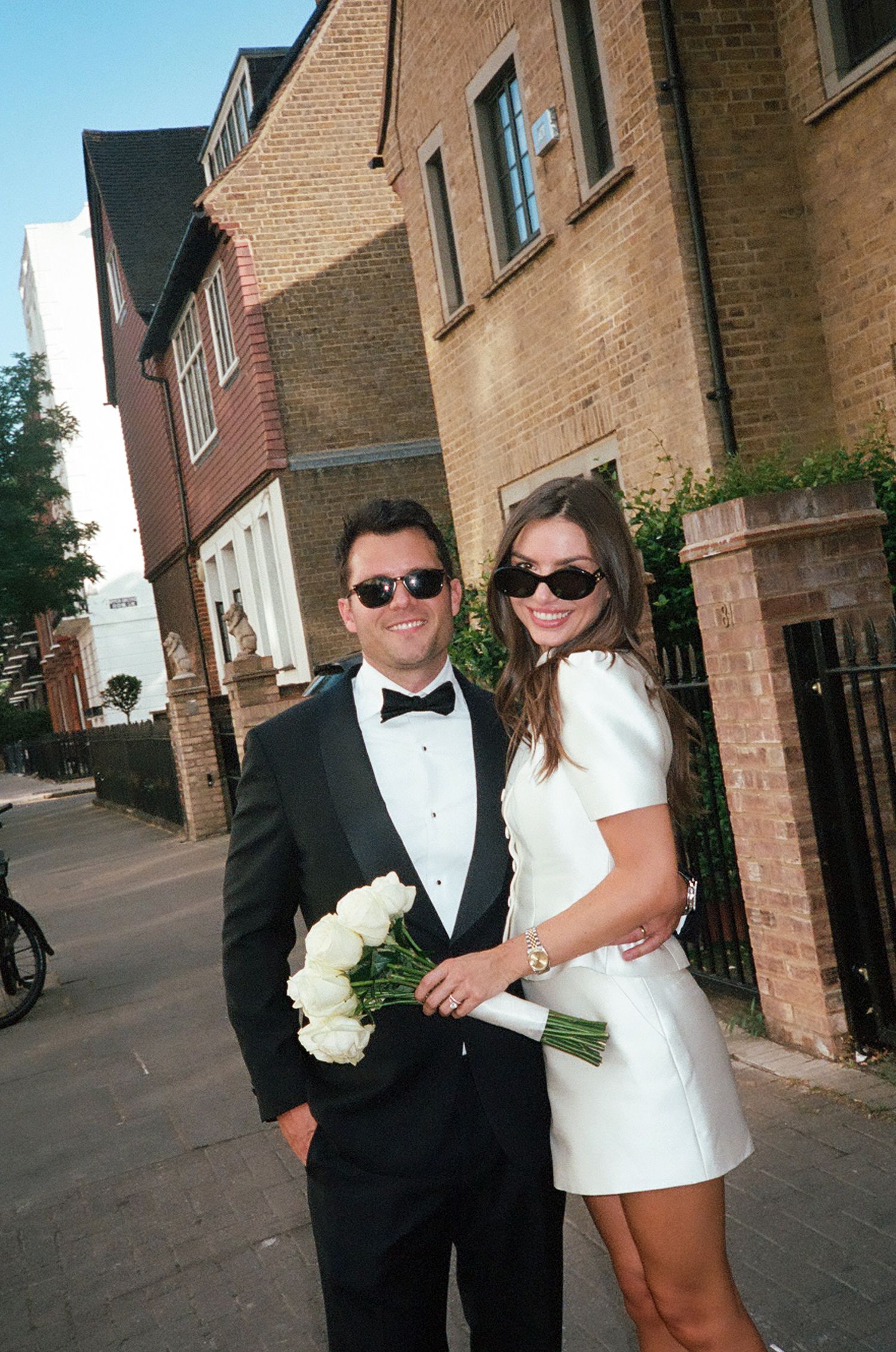 Smiling newlyweds in sunglasses posing with white bouquet on sunny London street.