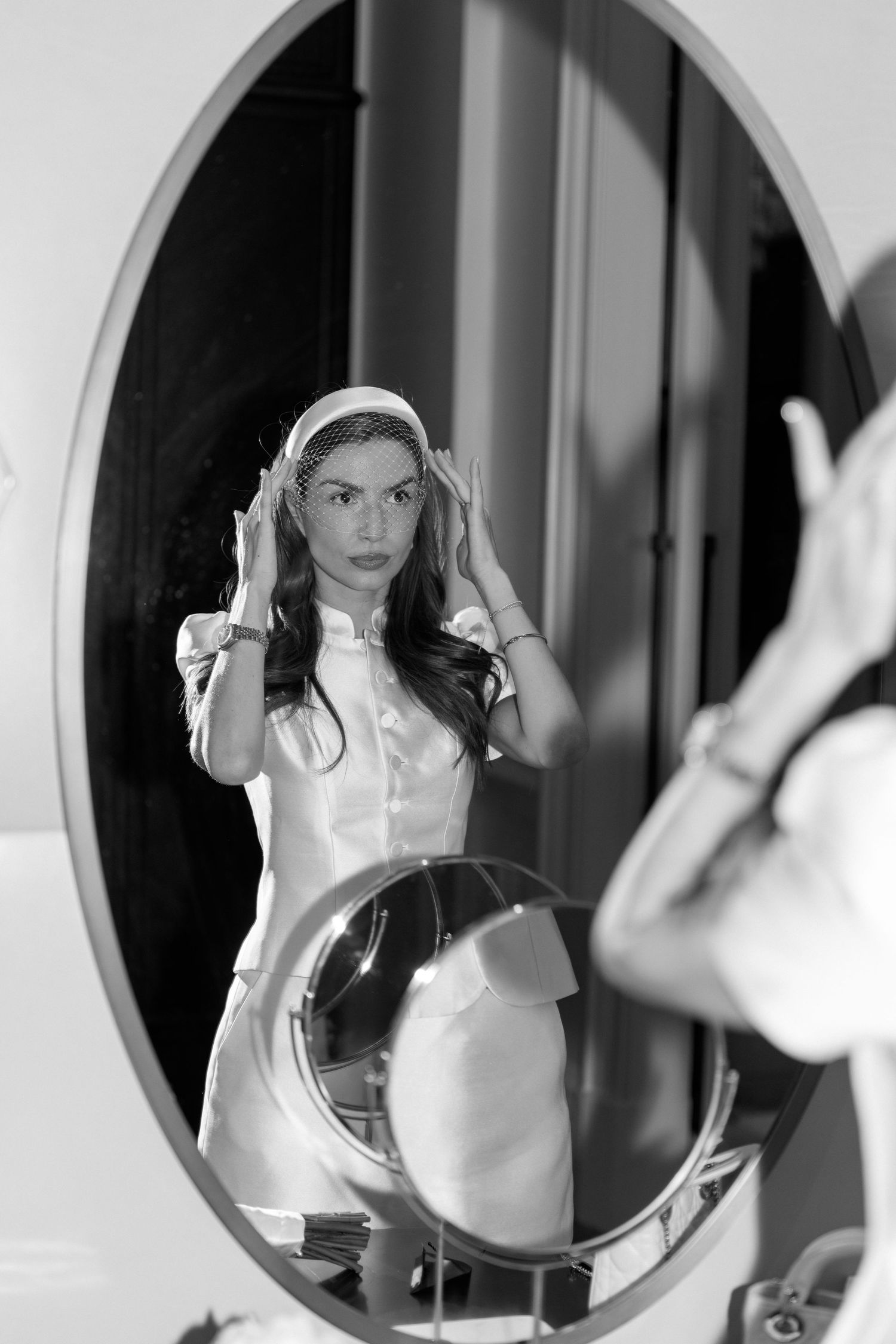 Bride in white mini dress adjusts veil while looking at her reflection in an oval mirror.