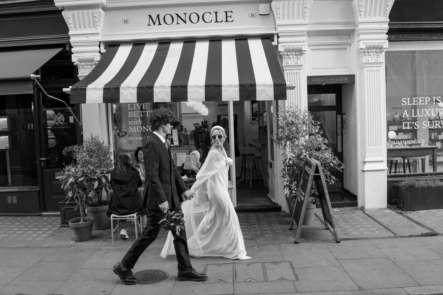 Couple in wedding attire walking past the Monocle storefront with classical columns.