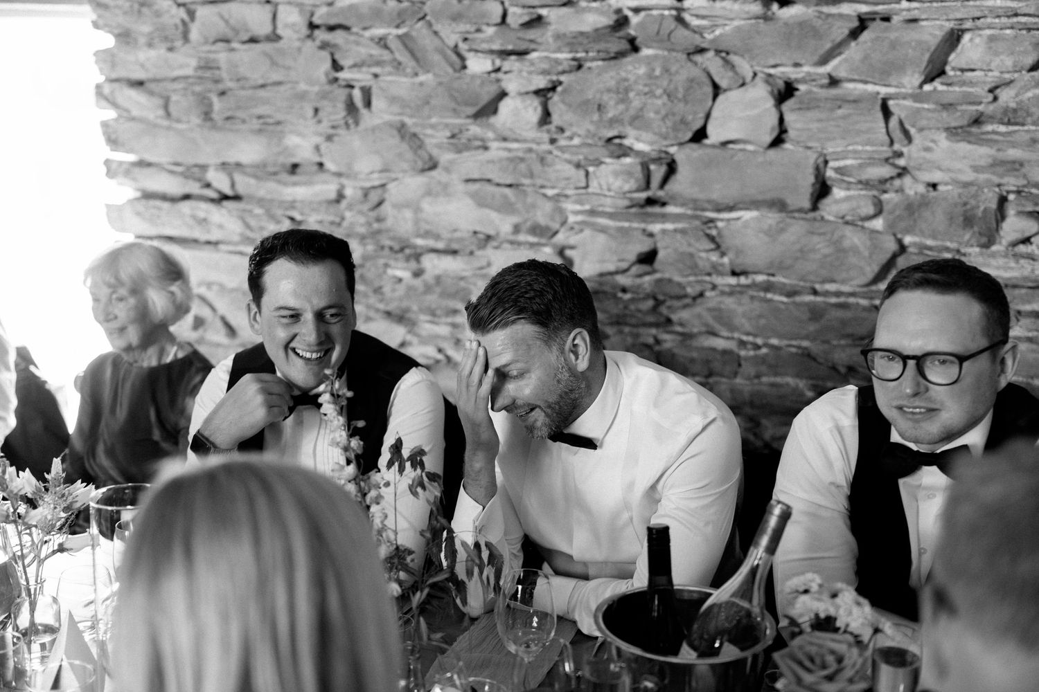 Wedding guests laughing and enjoying themselves at a reception with a rustic stone wall backdrop.