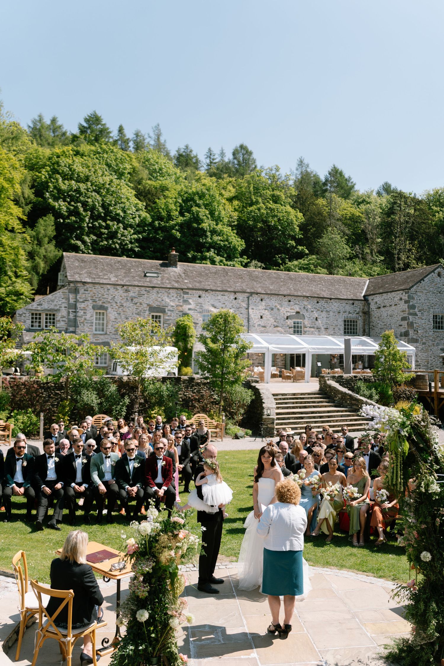 Outdoor wedding ceremony setup at a rustic stone venue with guests seated on wooden chairs and floral arch decorations.