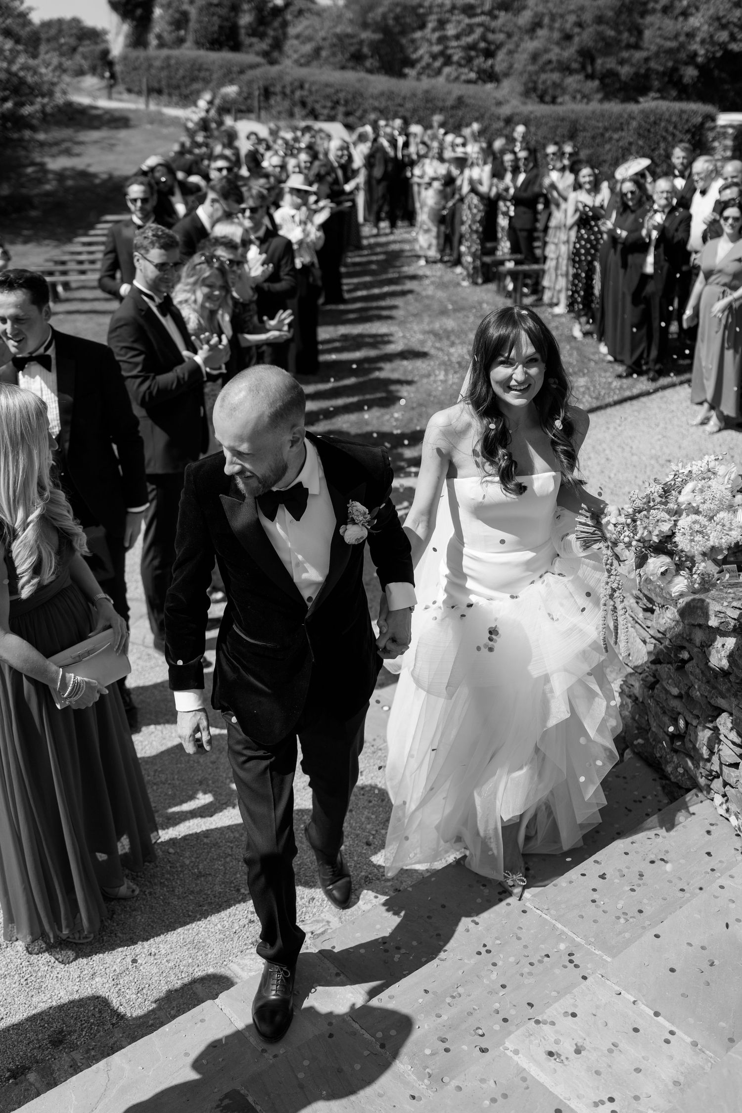 Black and white photo of a wedding procession walking through scattered confetti on a garden path.