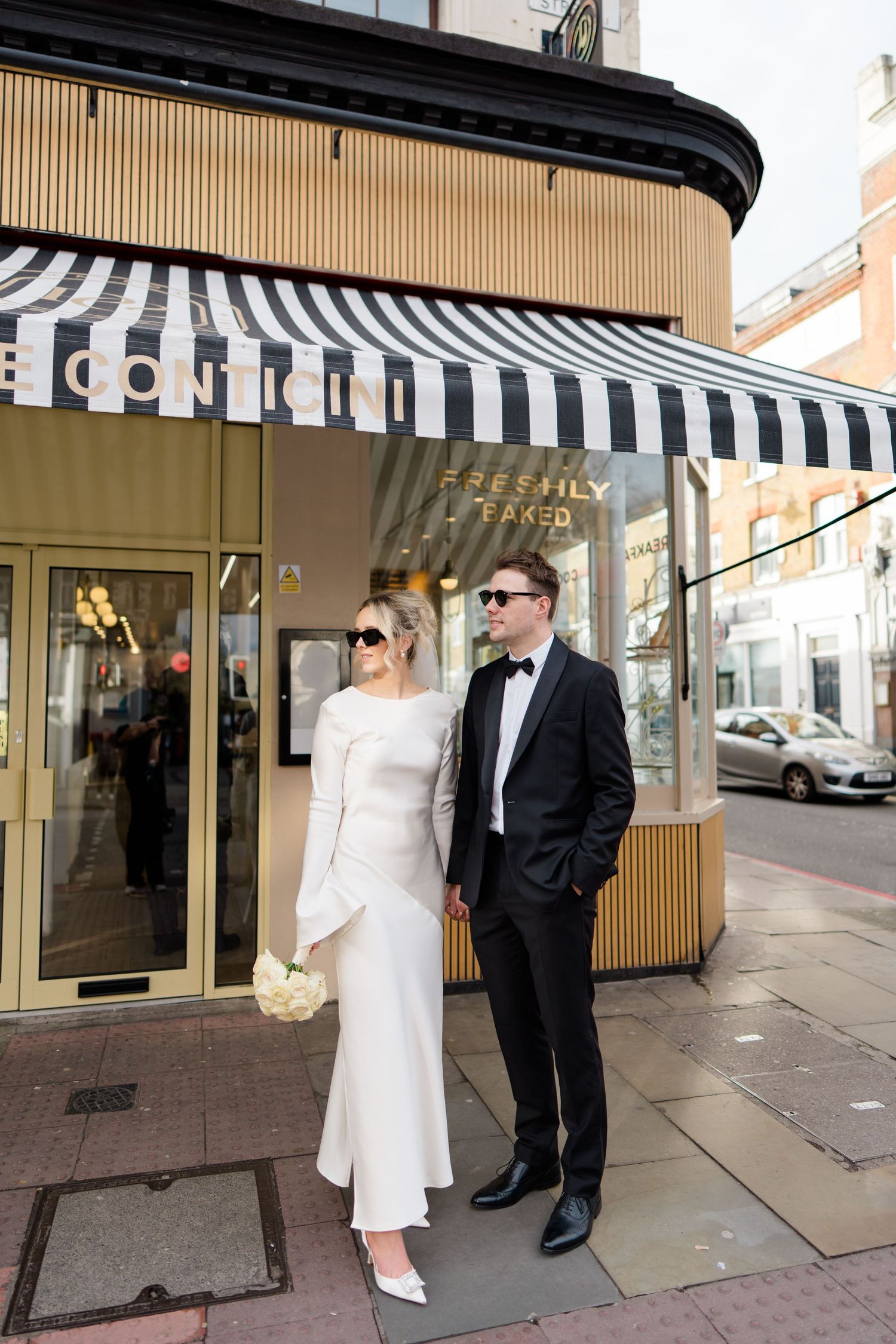 Stylish couple in wedding attire standing outside storefront with striped awning.