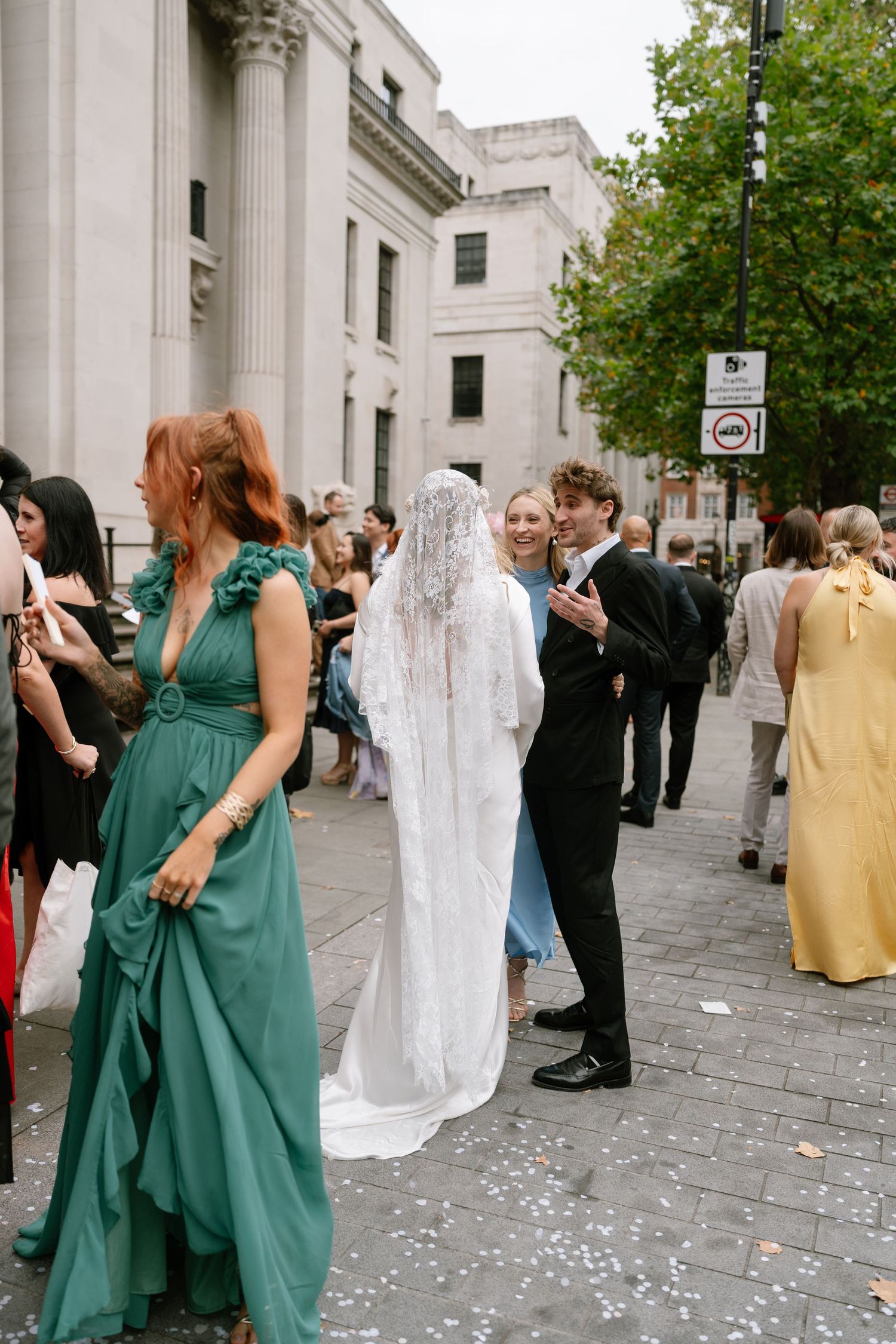 Wedding guests in green and white attire mingle outside a columned building.
