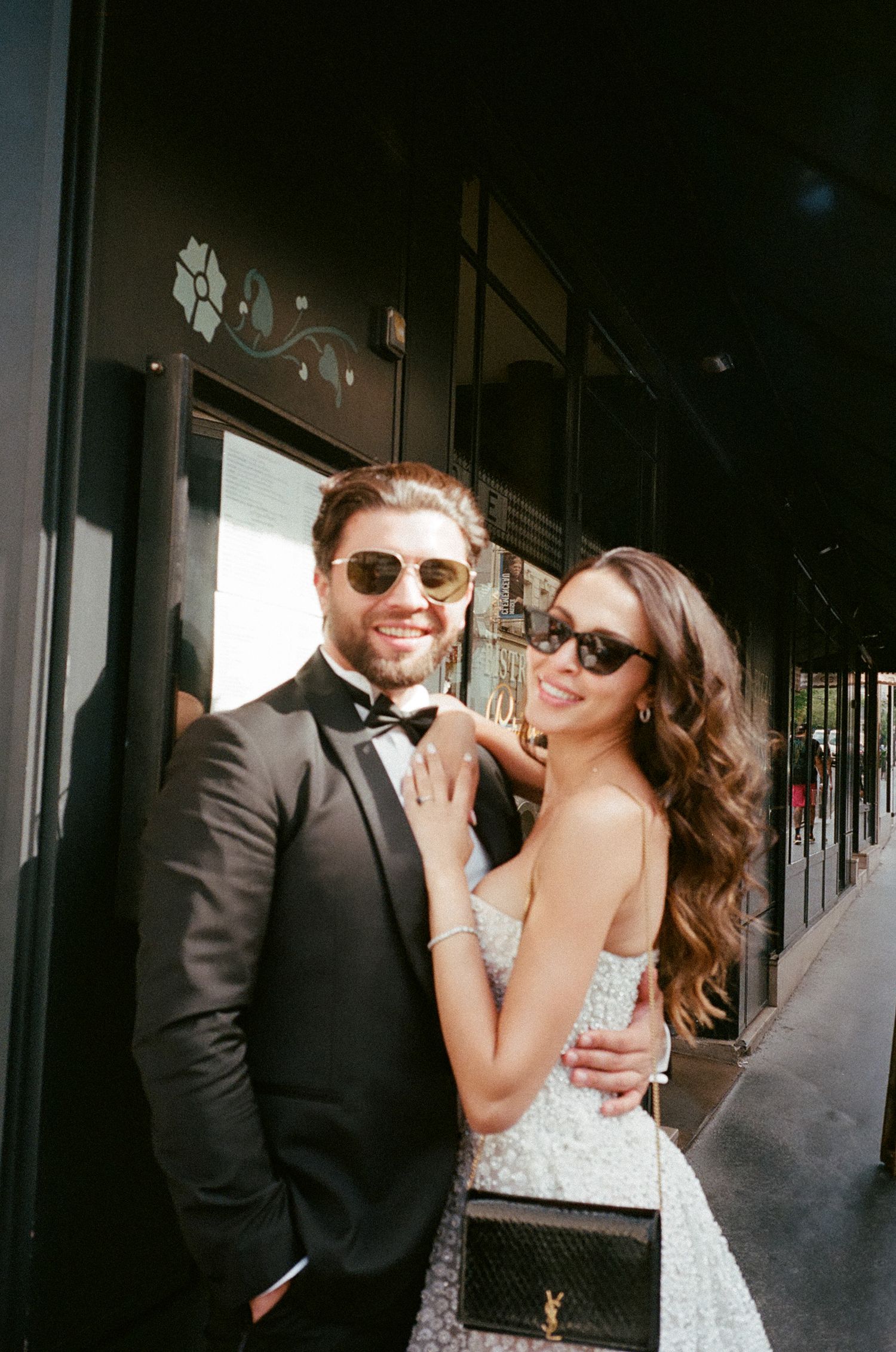 Stylish couple in formal attire and sunglasses pose together on a city street.