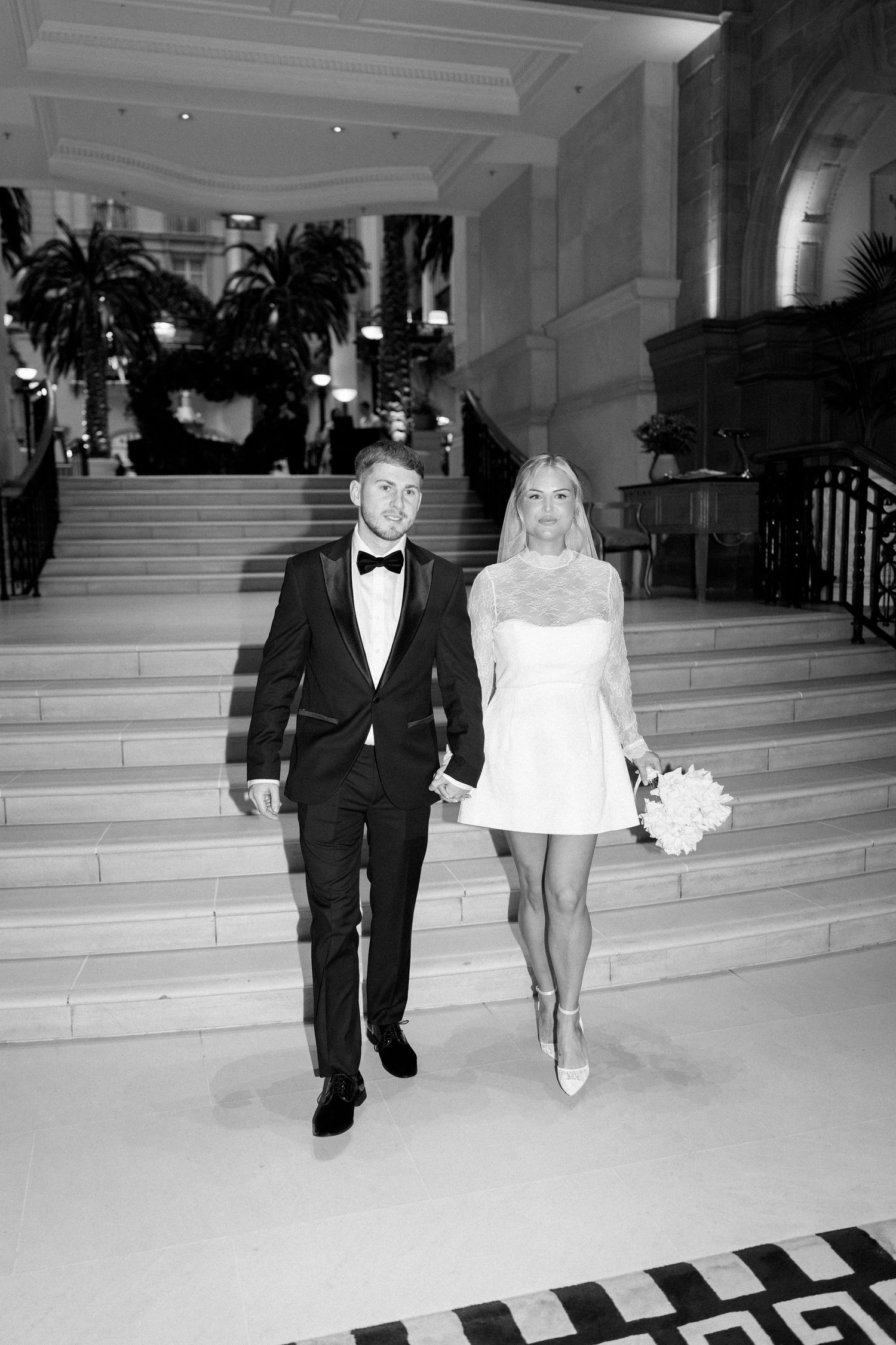 A couple in wedding attire walks down elegant marble stairs in a formal building interior.