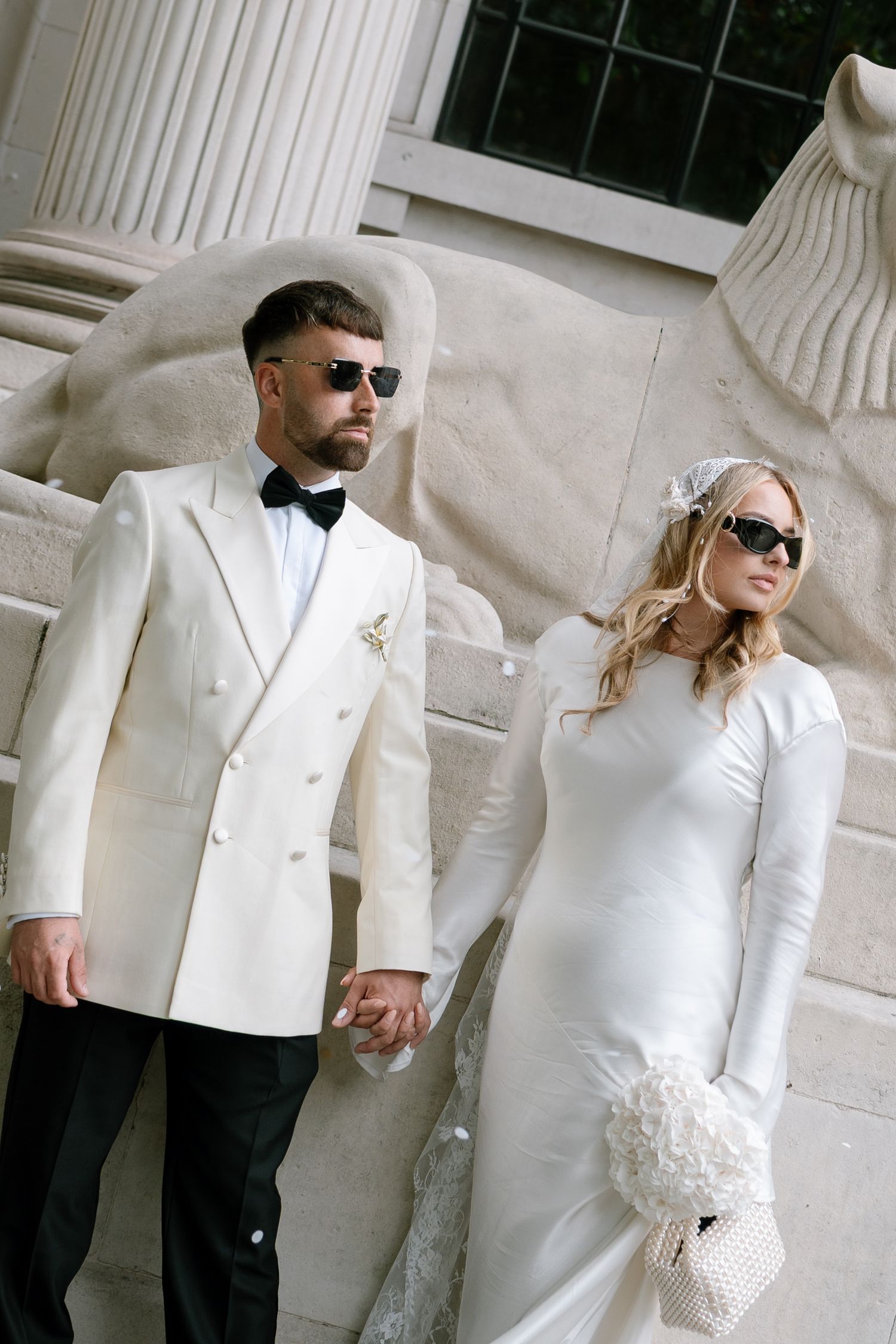 A stylish couple in white formal wear poses against a classical column.