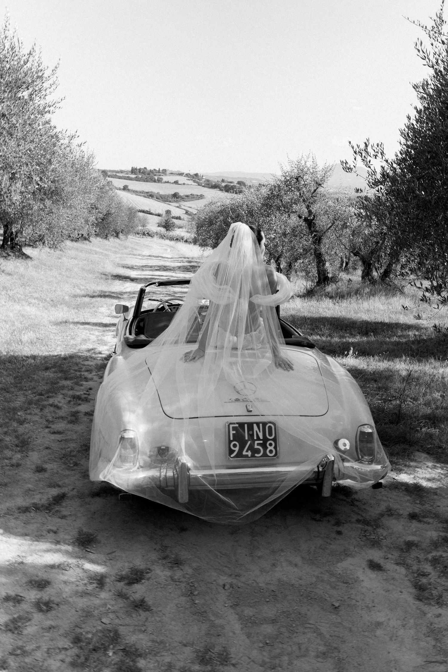 A vintage car decorated with wedding tulle drives through a scenic countryside path.