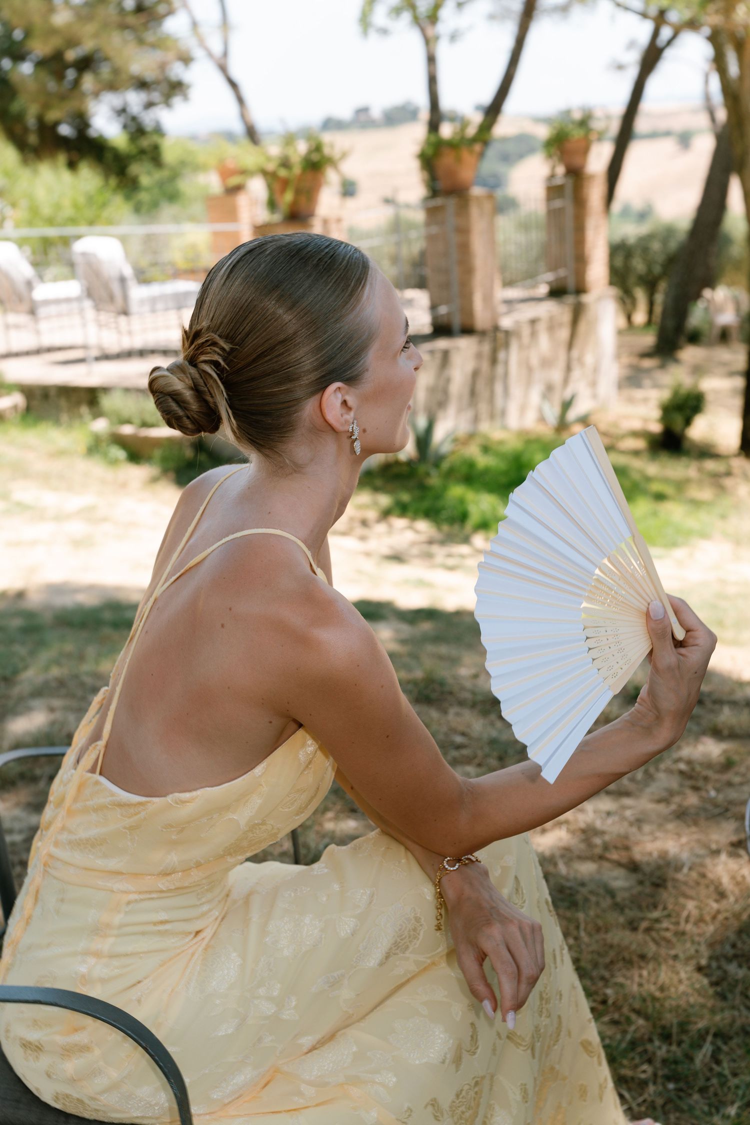 A person in a yellow dress sits outdoors holding a decorative hand fan.