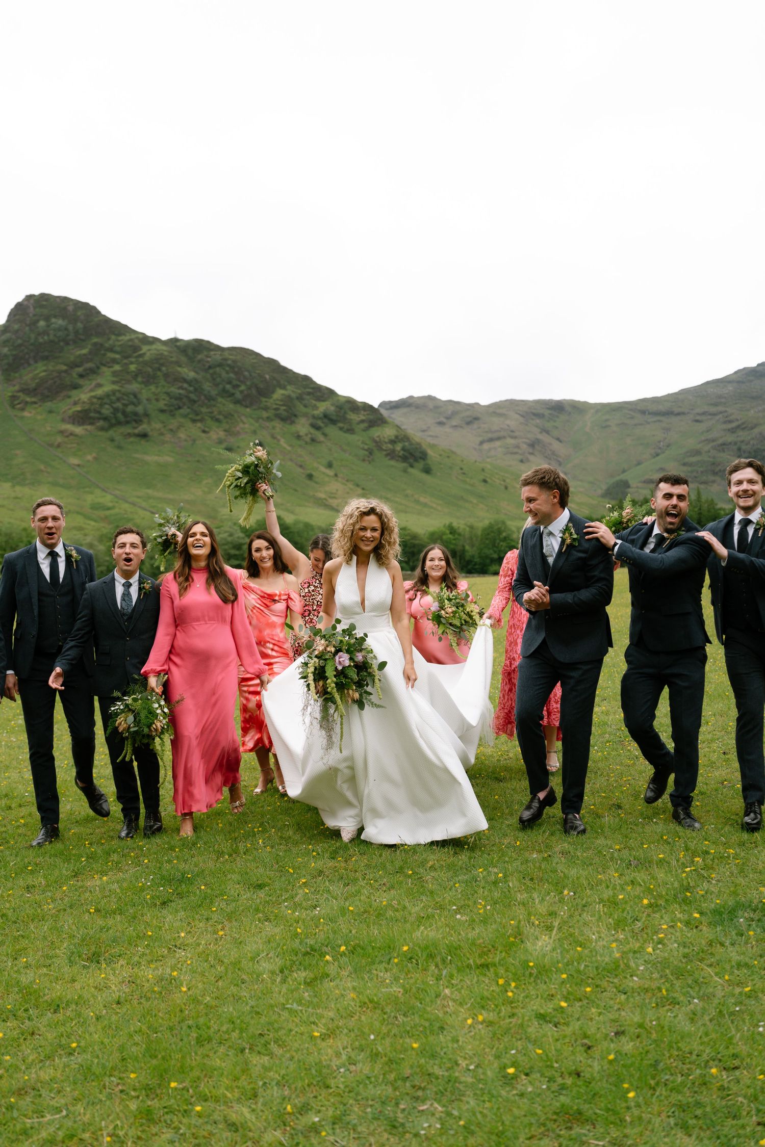Wedding party in pink dresses and black suits poses together on green hillside landscape.