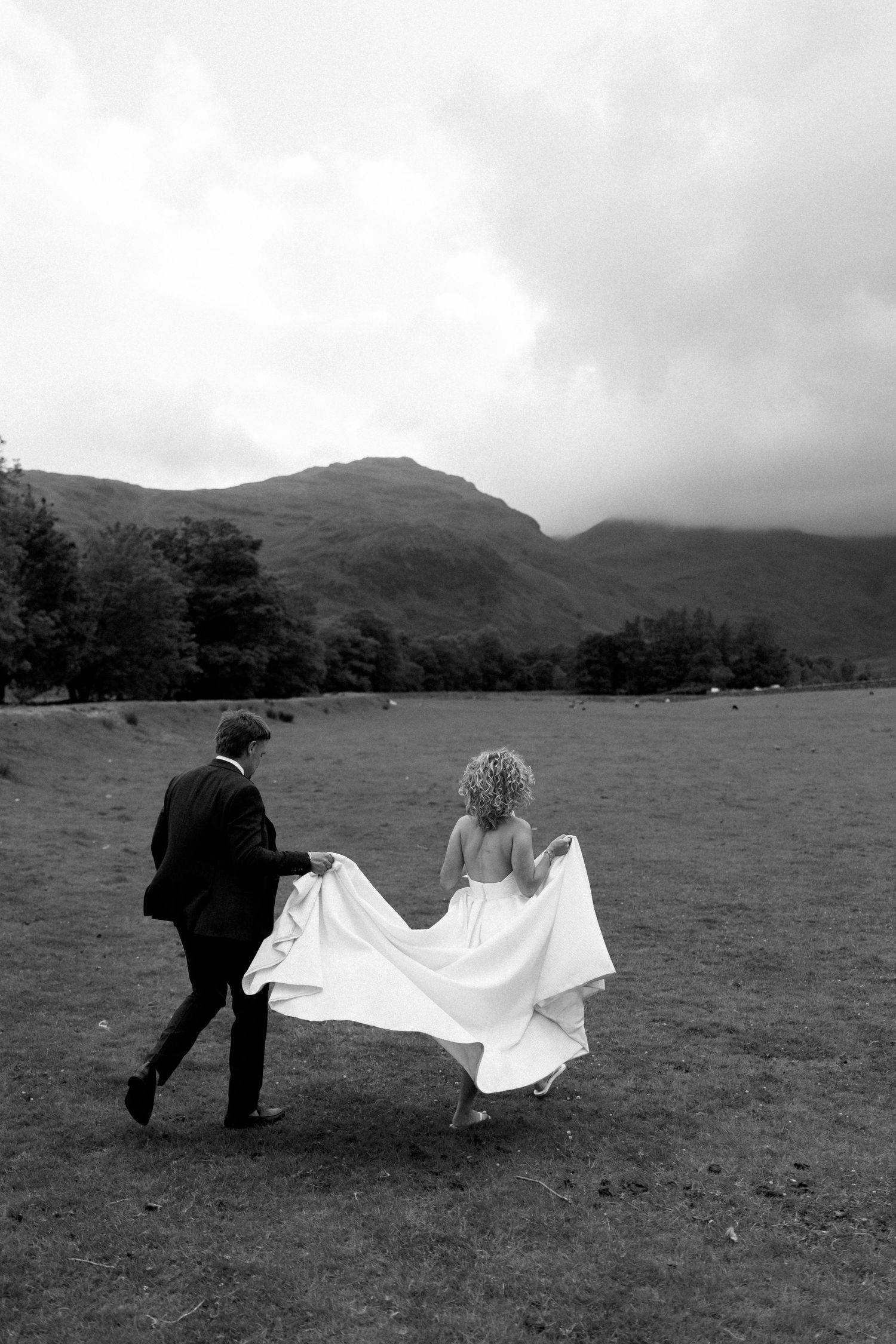 Wedding couple playfully interacting with dress in black and white against mountain landscape.