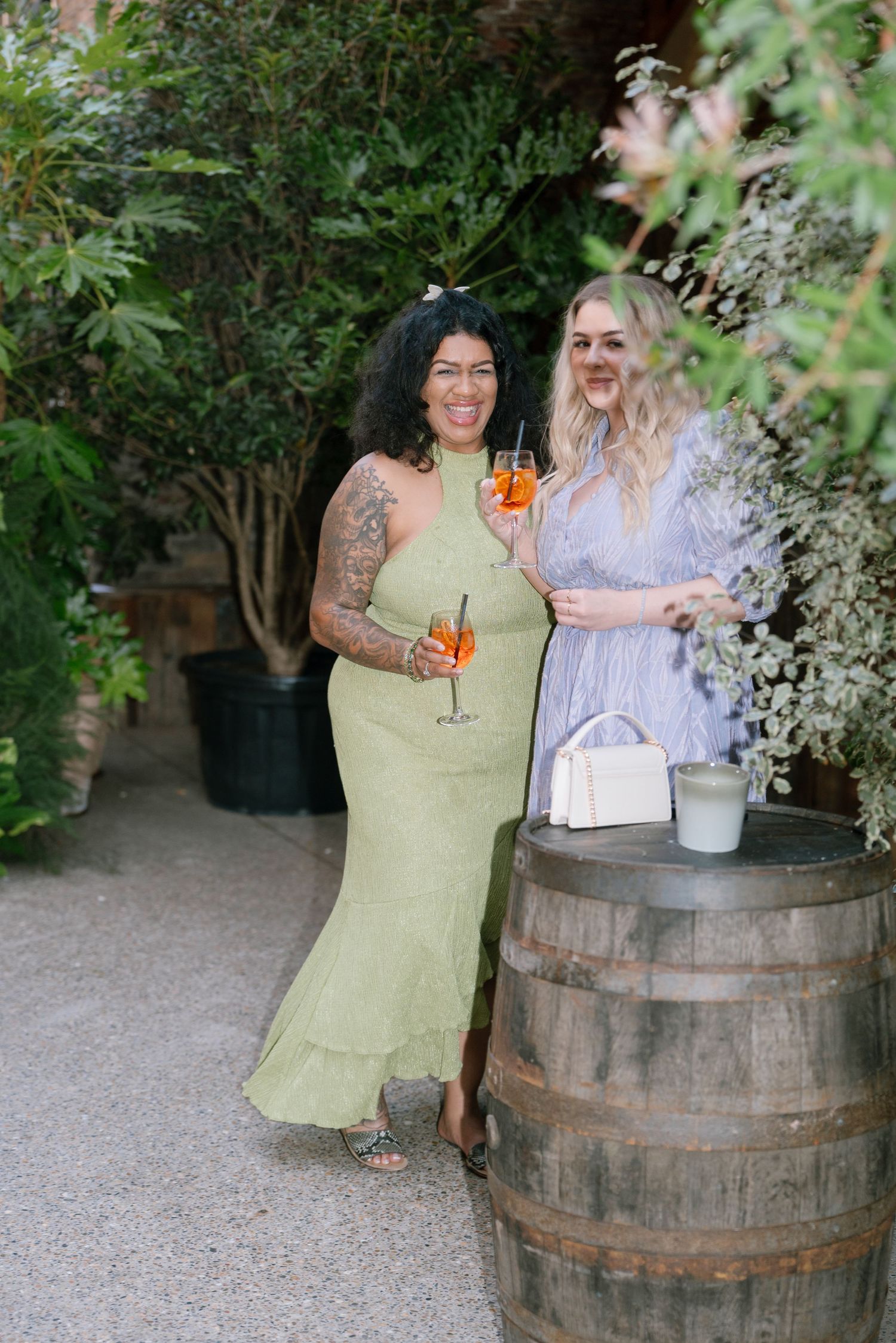 People enjoying cocktails in a garden setting with wooden barrels and greenery.