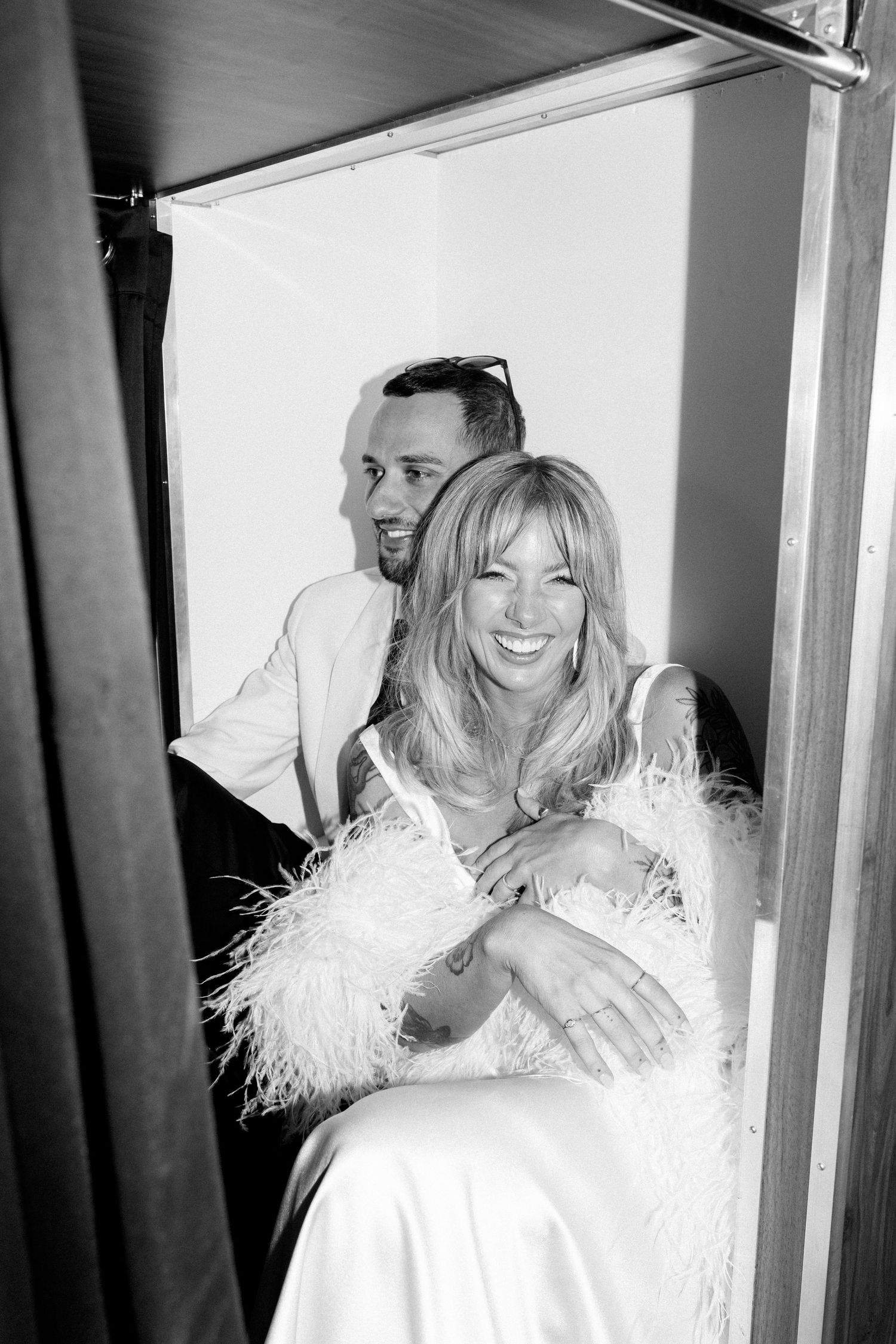 Black and white photo of a couple sharing a joyful moment in a white feathered wedding outfit.