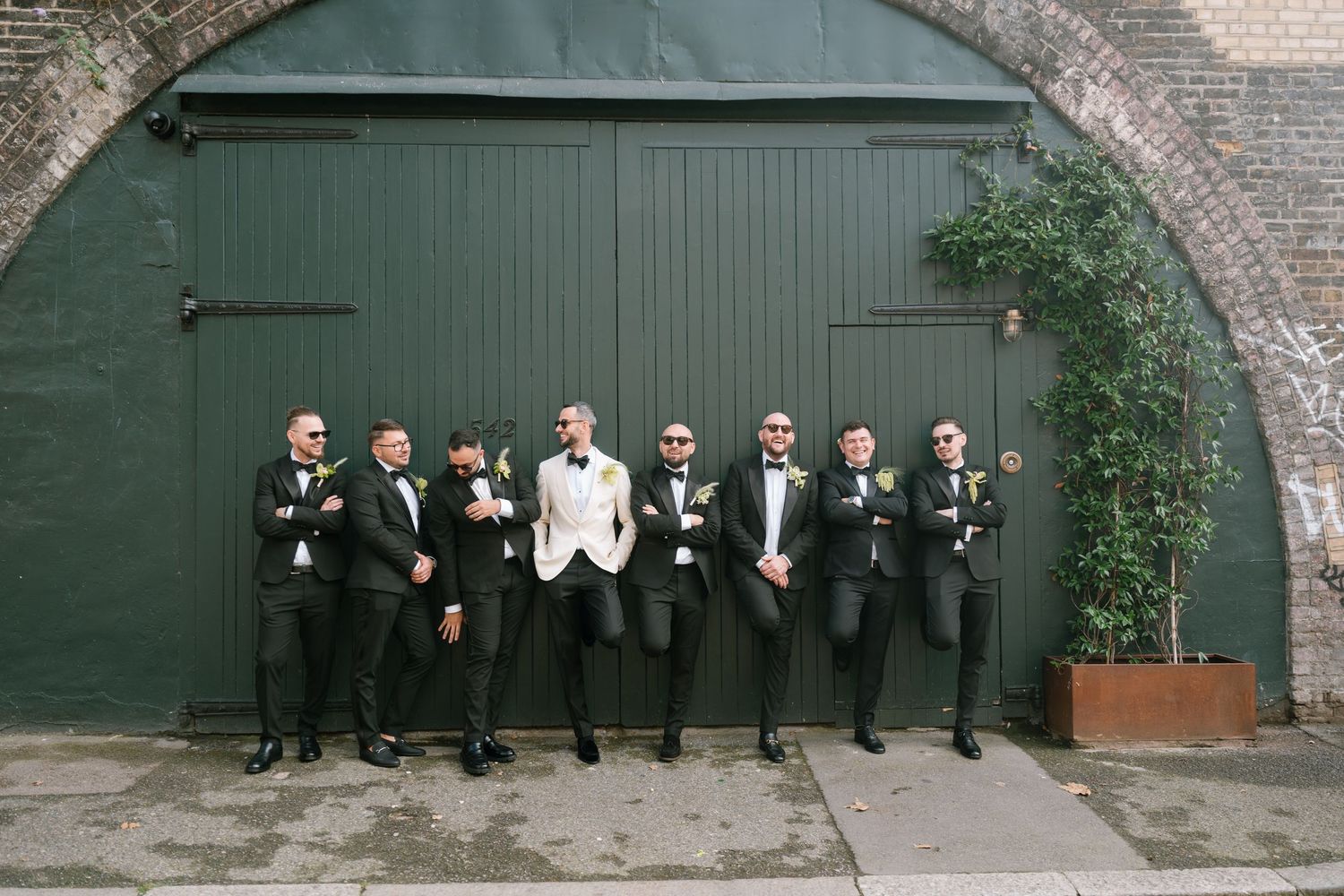 Group photo of wedding party members lined up against green doors and brick wall.
