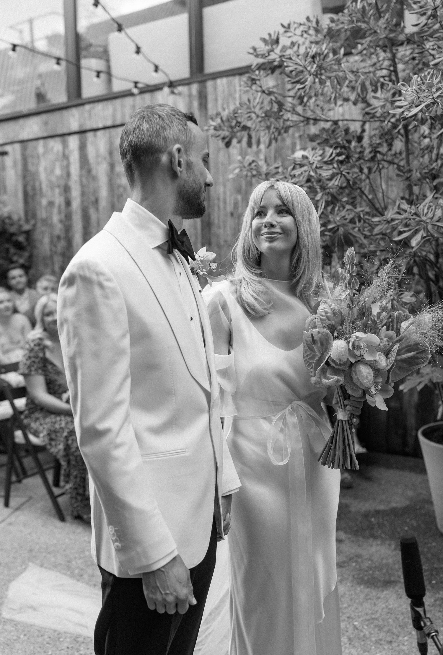 Elegant wedding couple in white attire sharing a candid moment together in an outdoor garden setting.