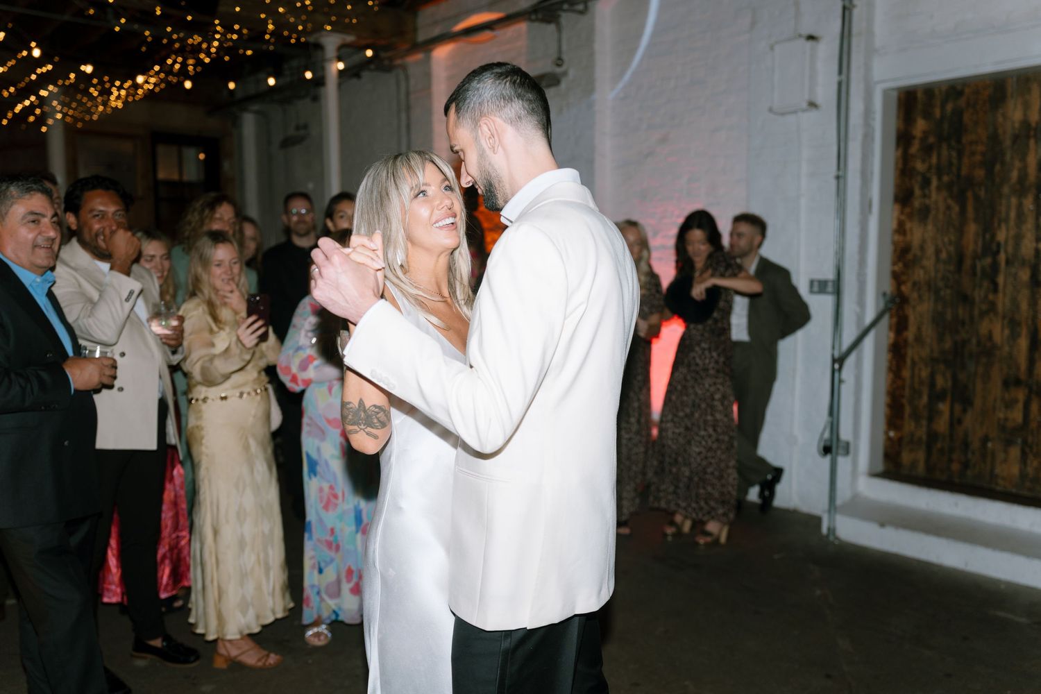 Guests dance and celebrate under warm string lights at an indoor wedding reception.