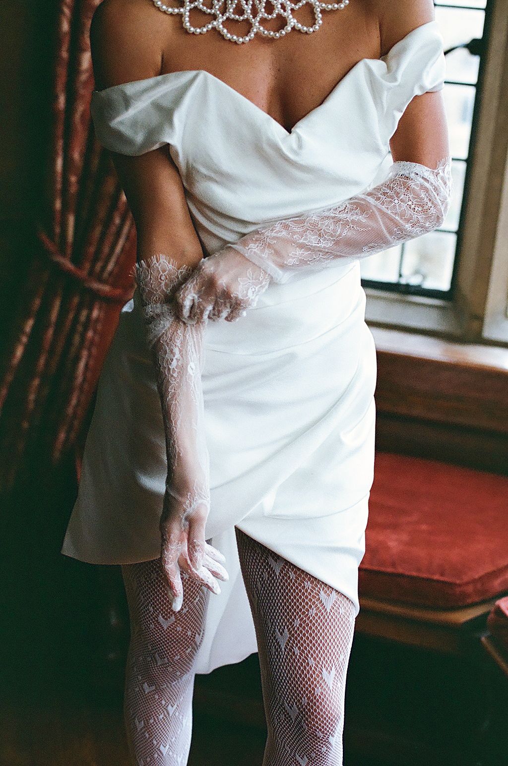 Elegant white bridal ensemble featuring delicate lace gloves and fishnet stockings against a dark interior setting.