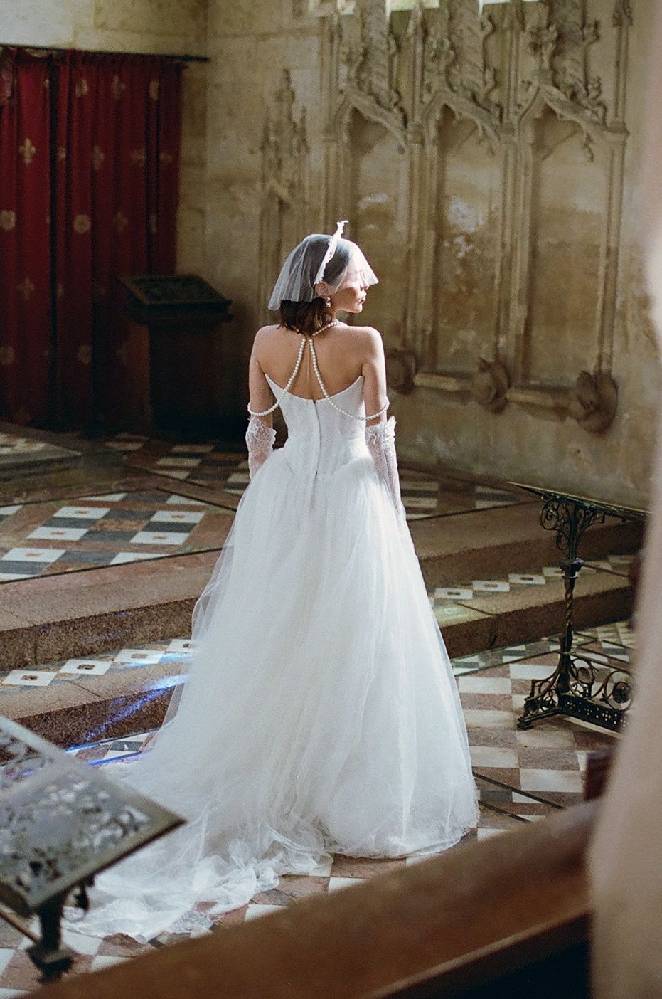 A bride in an elegant white wedding gown stands in a historic church with ornate gothic architecture and checkerboard floors.
