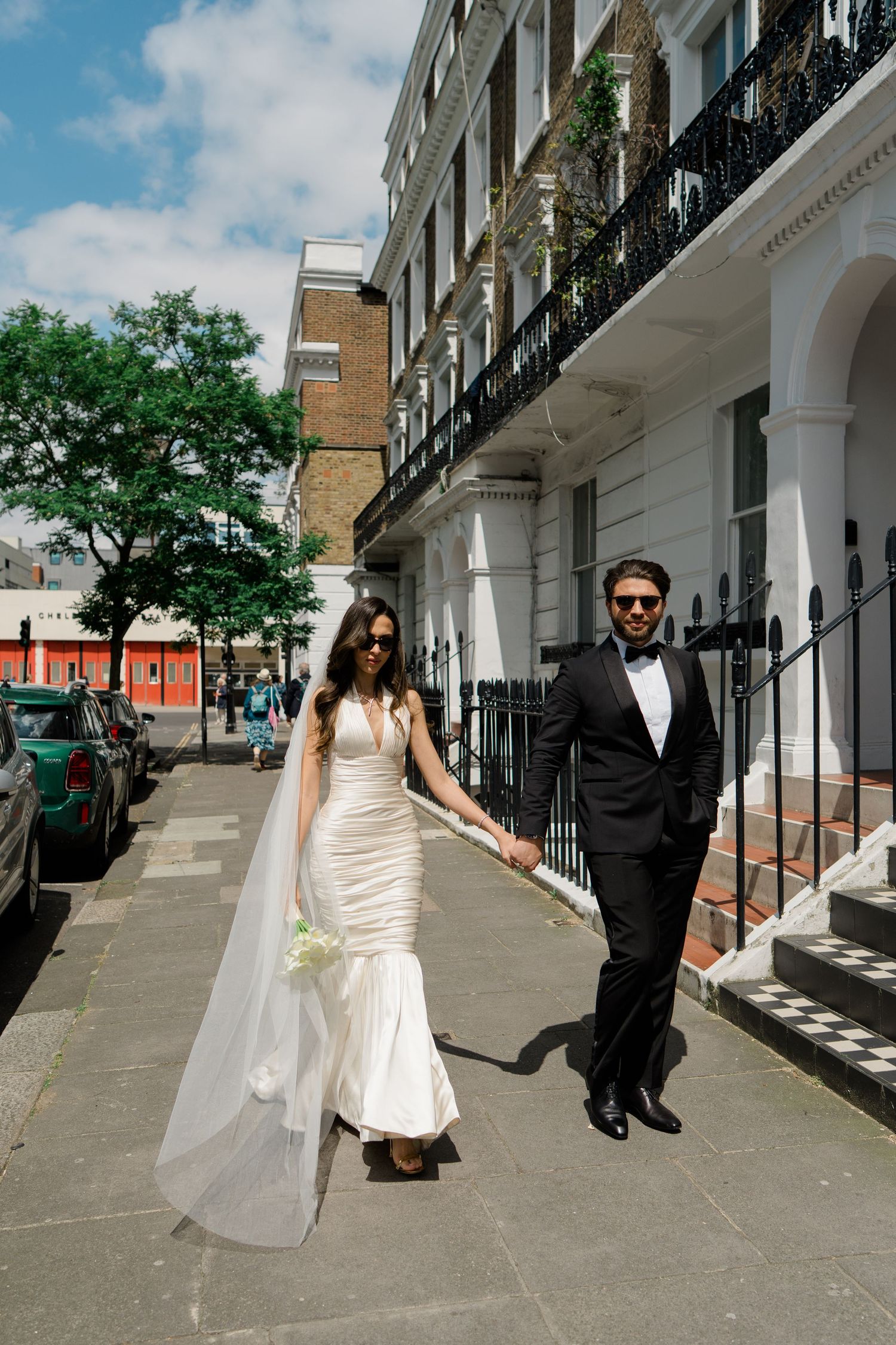 Bride in white gown and groom in black tuxedo holding hands walking on a sunny London street.