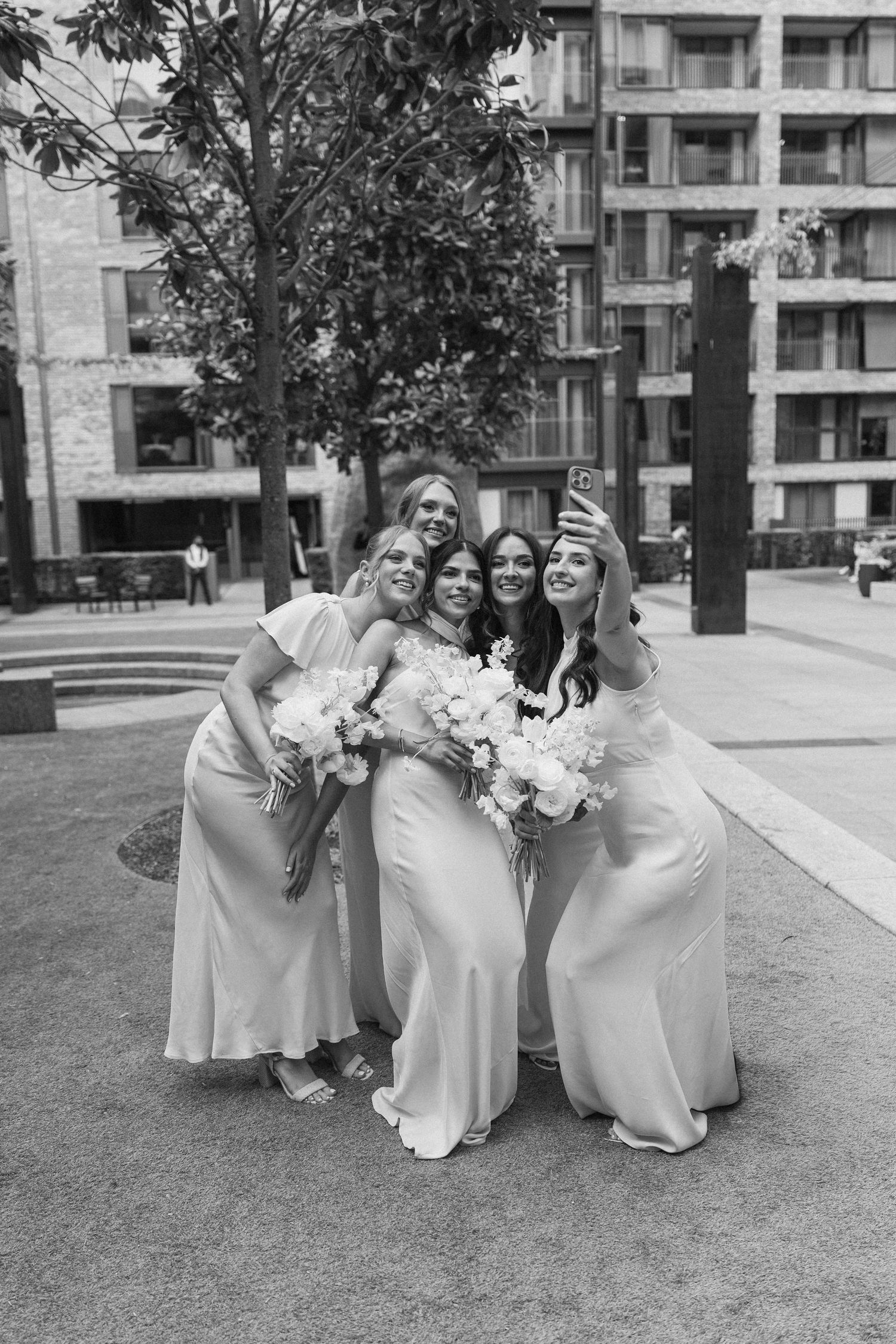 Bride with three bridesmaids taking selfie outdoors holding white bouquets in matching satin gowns.