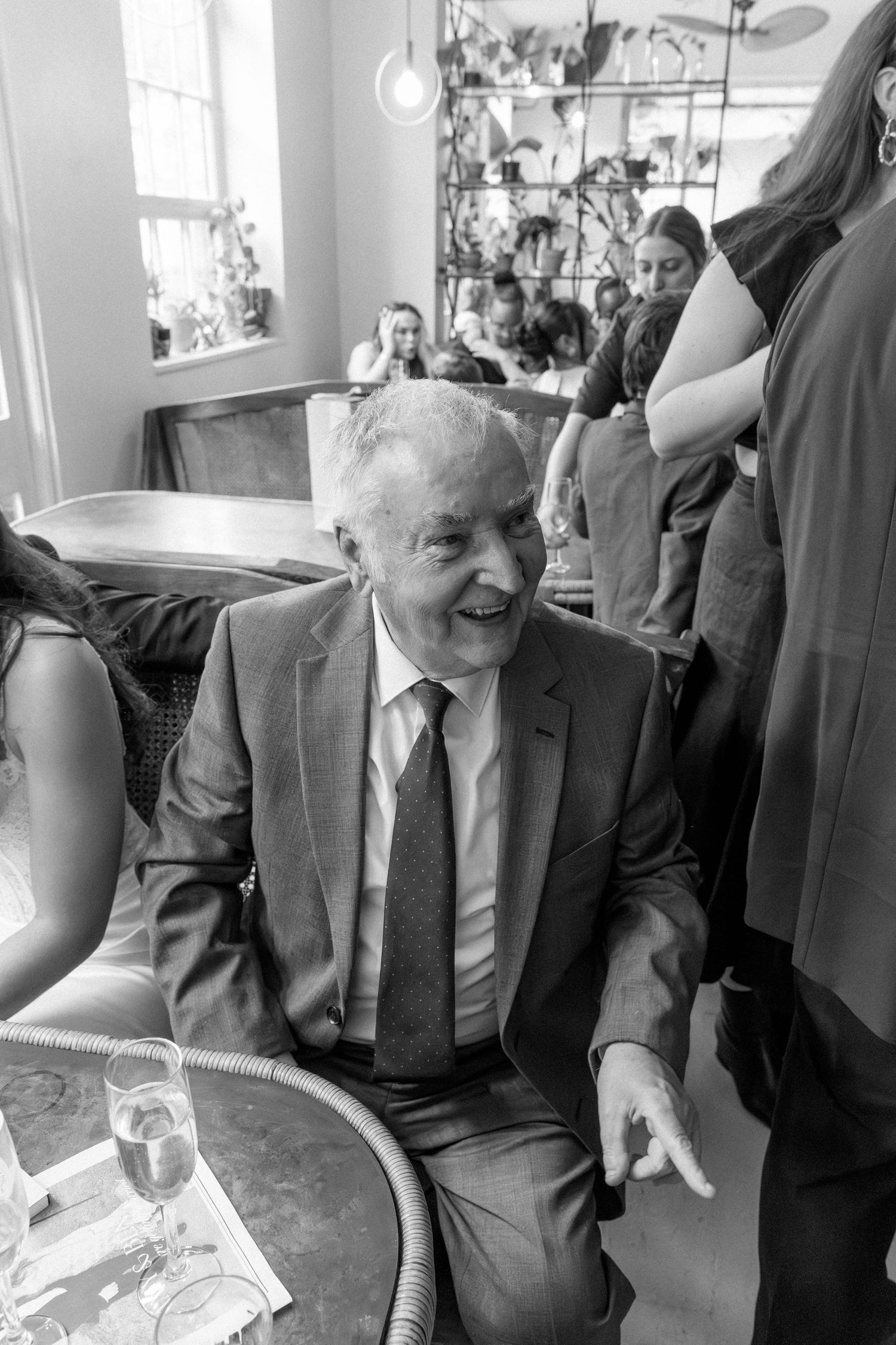 Elderly man in suit and tie laughs joyfully while seated at a table in a busy restaurant.