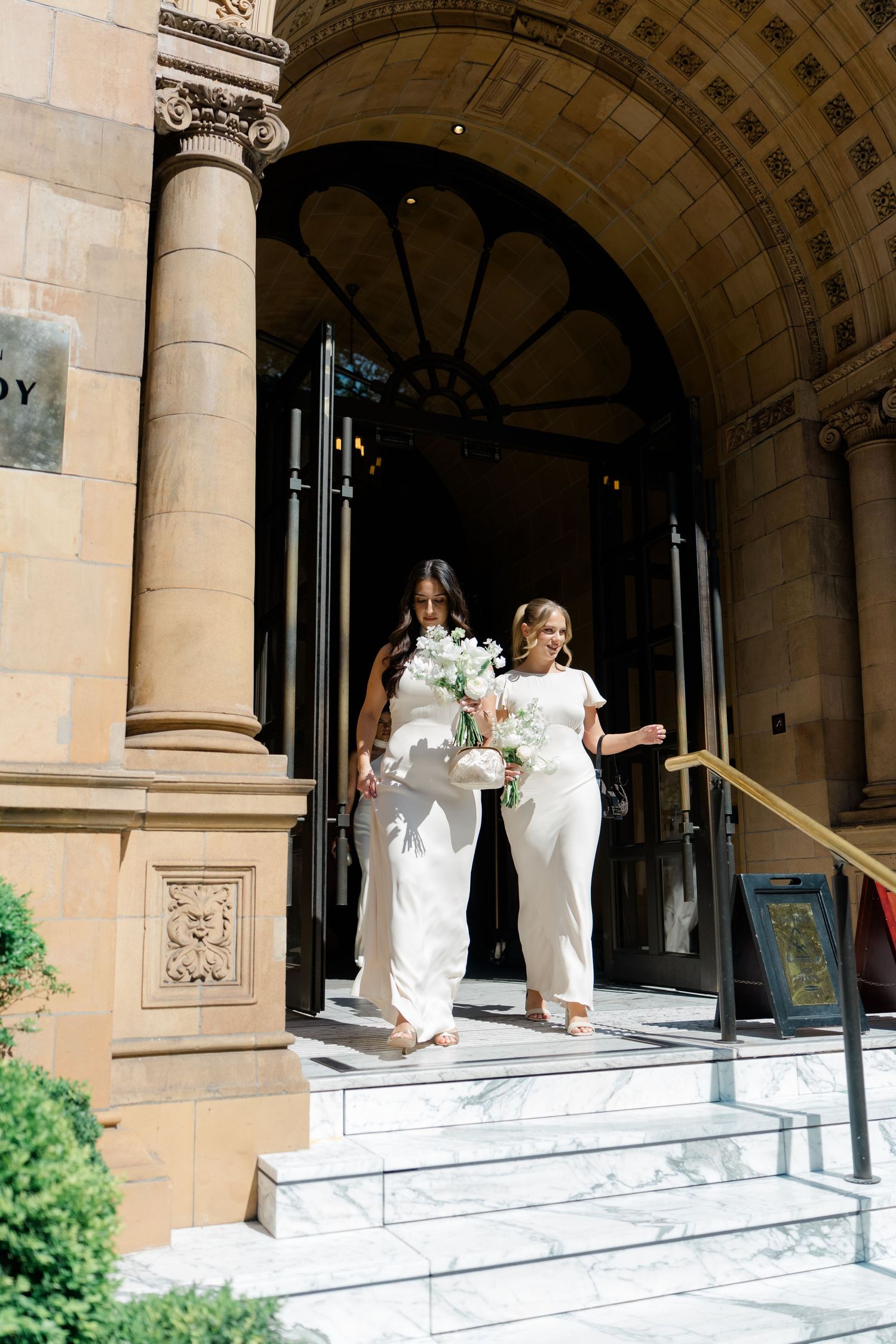 Two brides in white gowns carrying bouquets descend marble steps outside an elegant stone building.