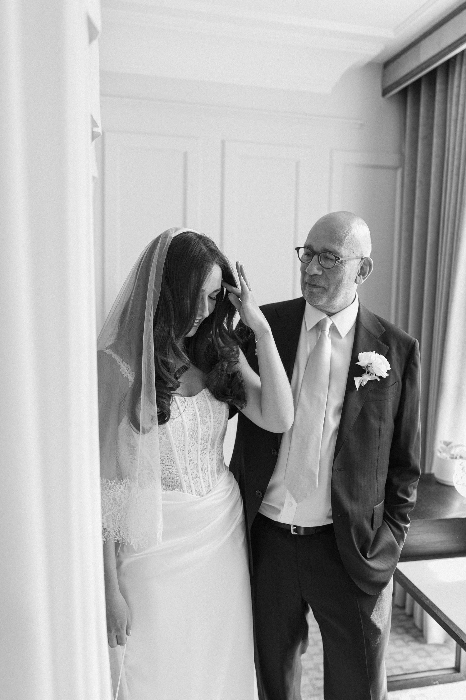 Black and white photo of a bride adjusting her veil while her father looks on lovingly.