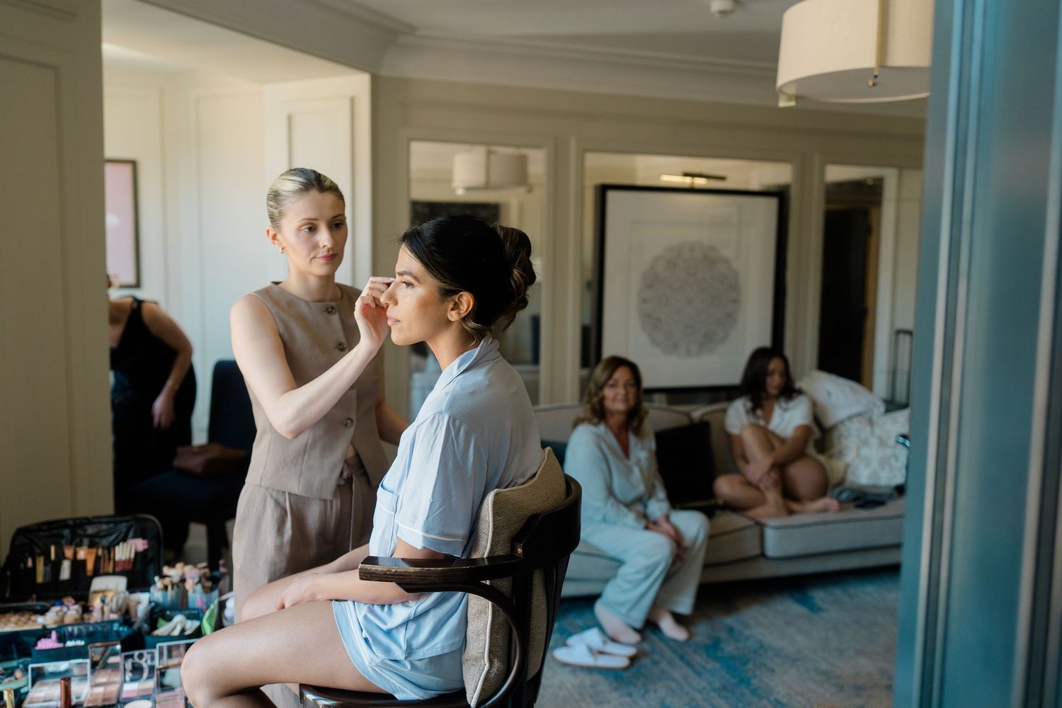 A makeup artist applies cosmetics to a seated bride while bridesmaids relax in the background.