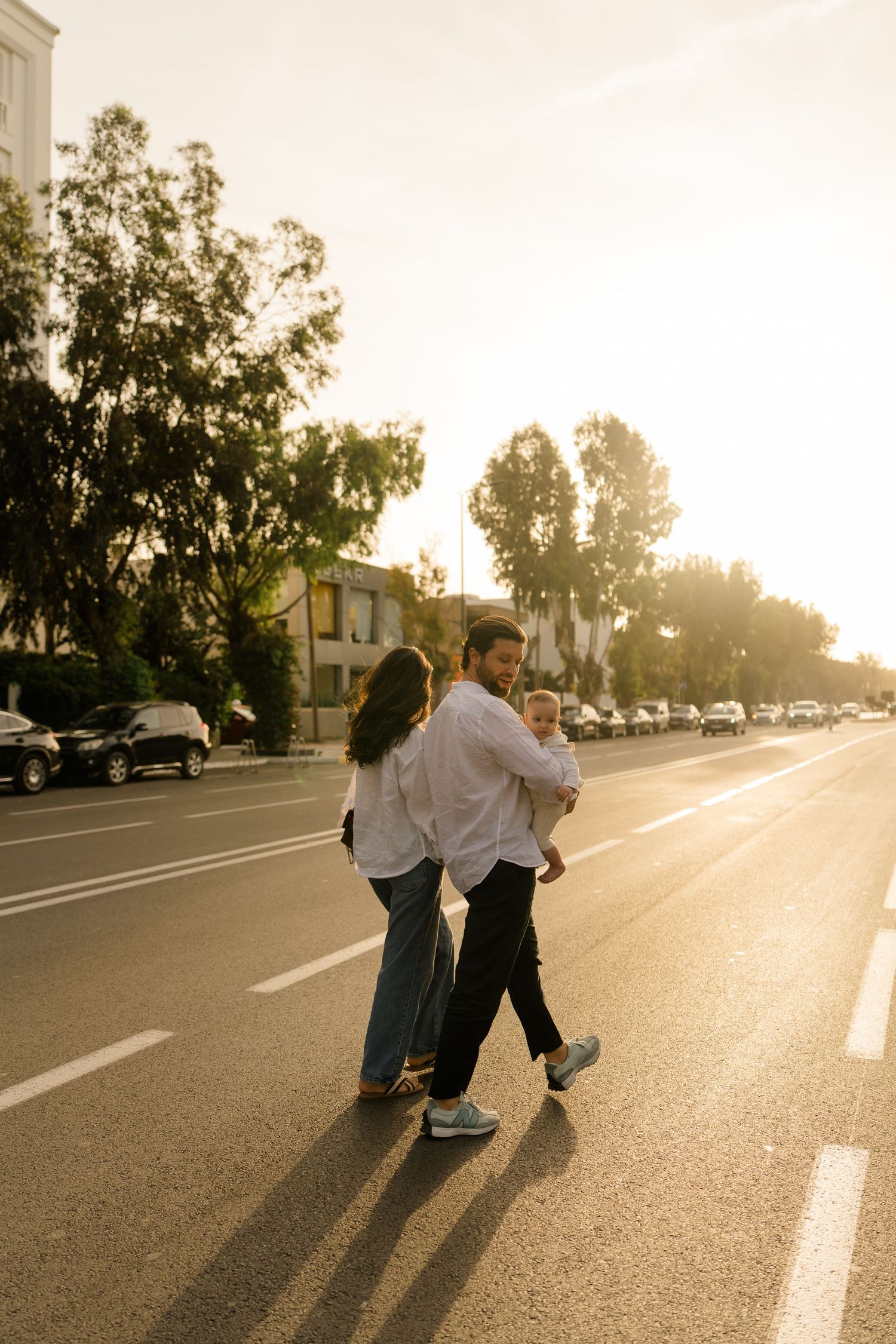 Family crossing a sunlit street at golden hour, with trees and parked cars in the background.