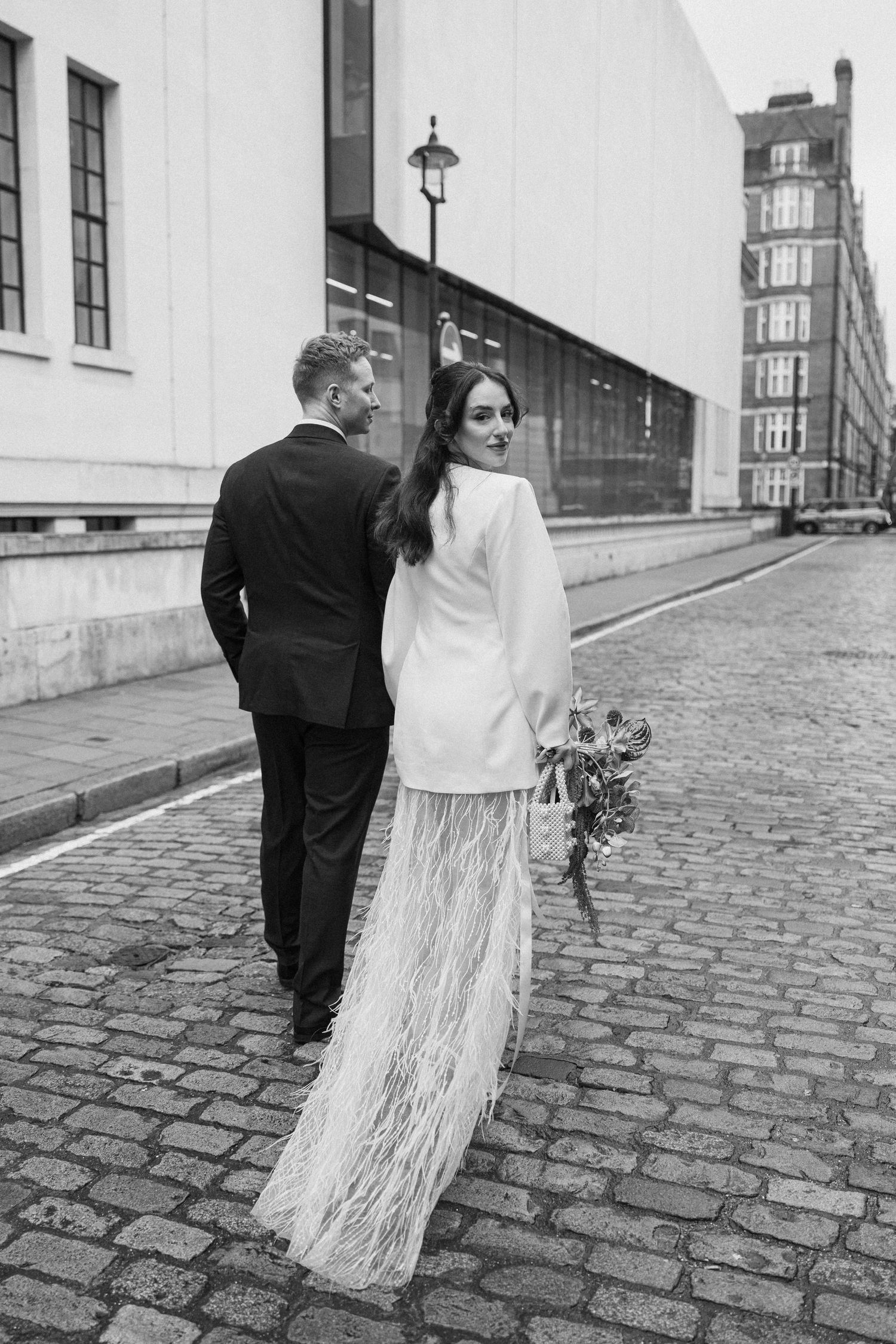 Black and white photo of bride in white feathered skirt and groom walking on cobblestone London street.