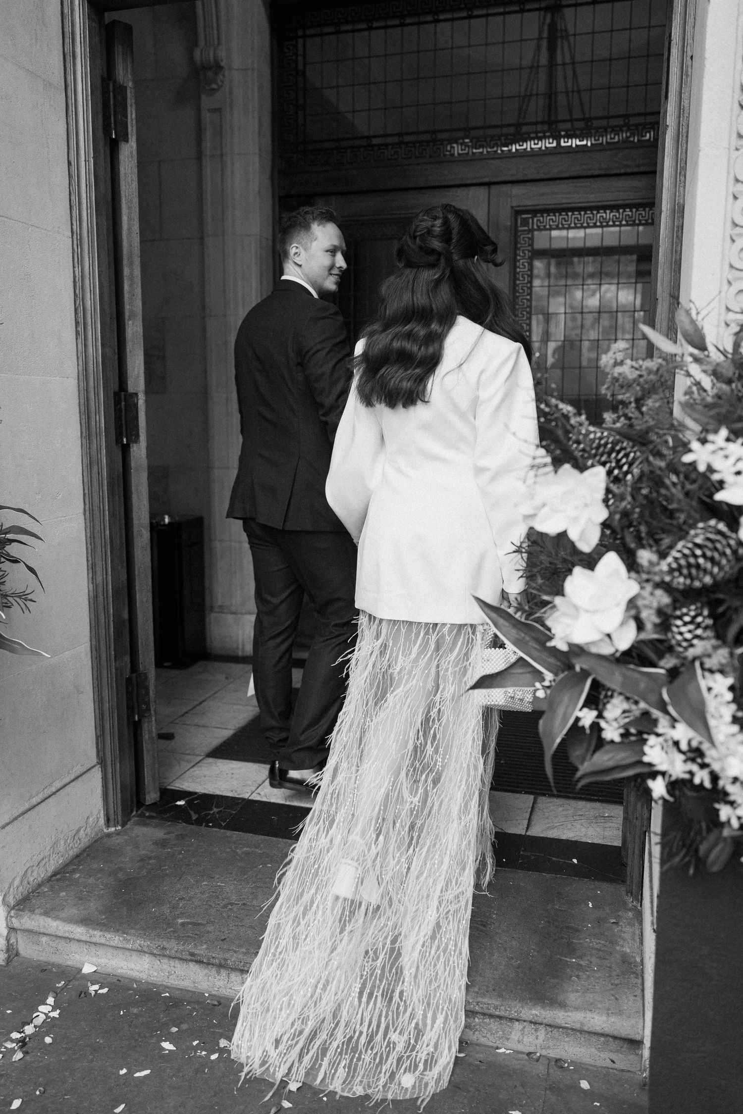 Black and white photo of bride in feather skirt and groom entering a building.