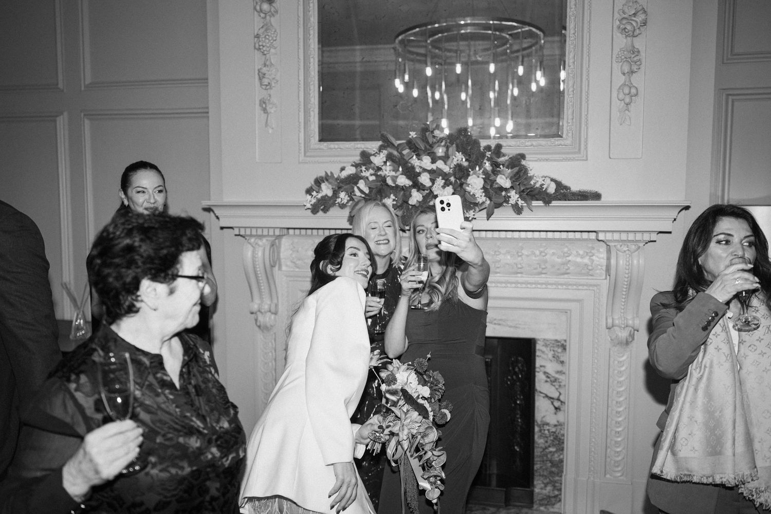 Black and white photo of bride greeting guests near ornate fireplace at wedding reception.