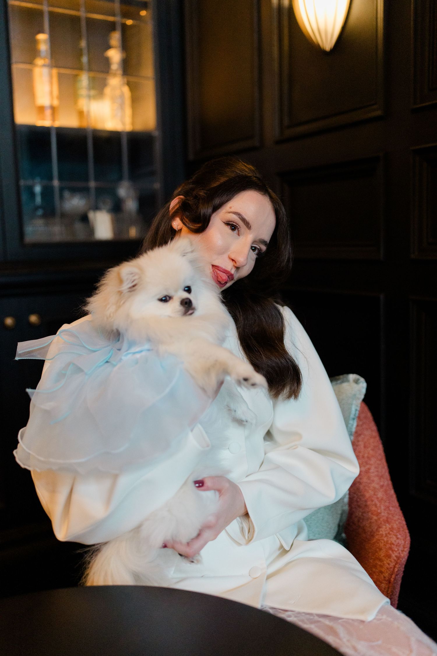 Bride in white robe holding fluffy Pomeranian dog with blue bow in elegant dark interior.