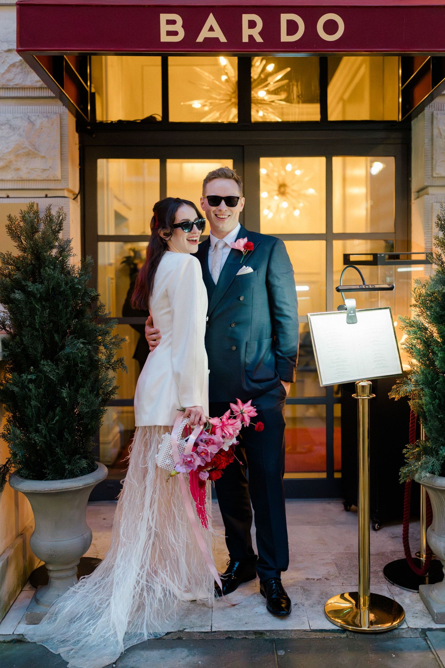 Bride and groom in sunglasses pose outside Bardo restaurant, bride holding pink bouquet, groom in navy suit.