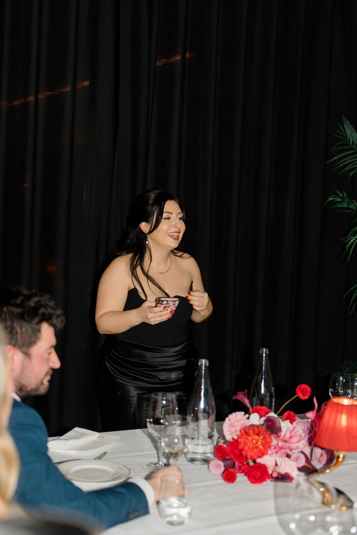 Woman in black dress giving speech at wedding reception dinner with floral centerpiece.