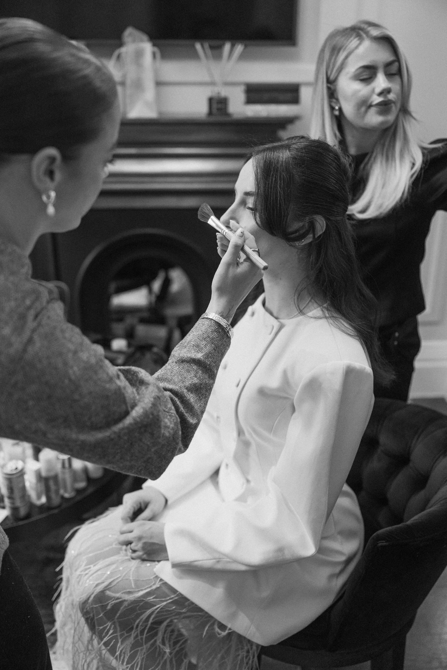 Black and white photo of bride having makeup applied by artist with hairstylist in background.