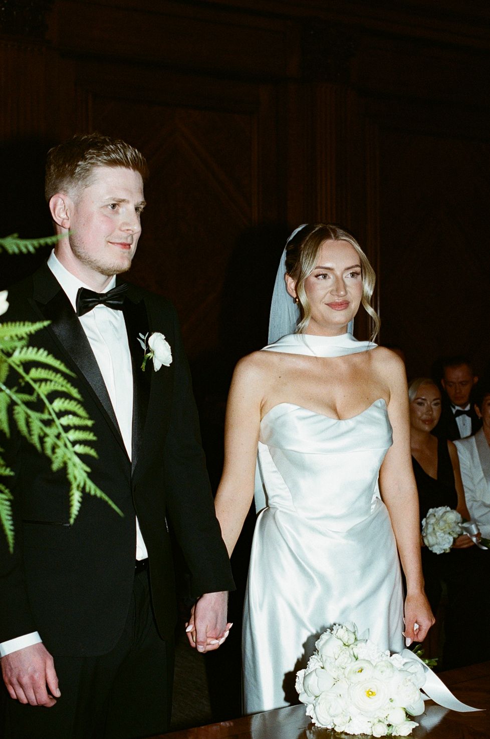 Bride in white strapless gown with veil and groom in black tuxedo holding hands at wedding ceremony.