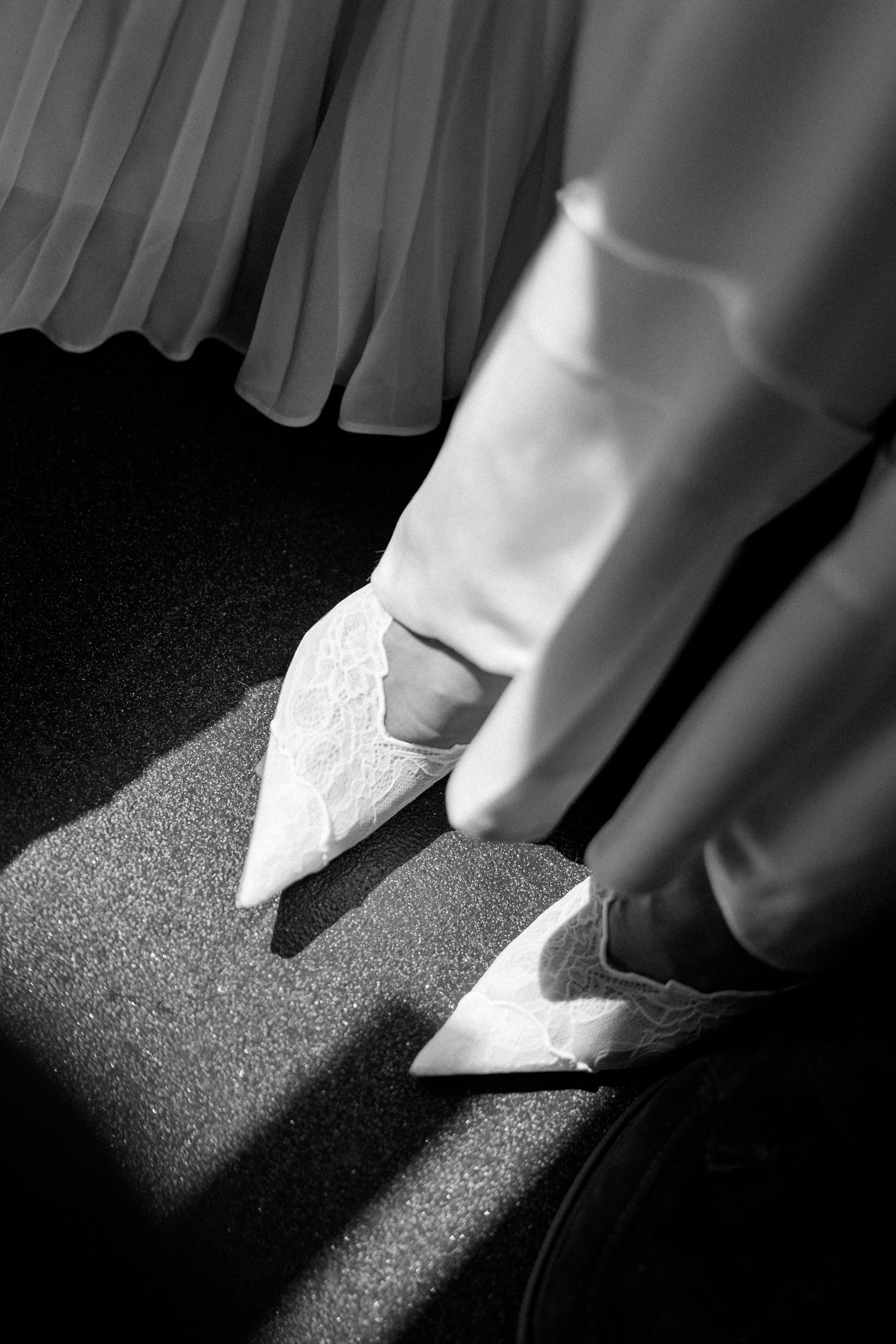 Close-up black and white photo of a bride wearing elegant white lace pointed-toe heels on her wedding day.