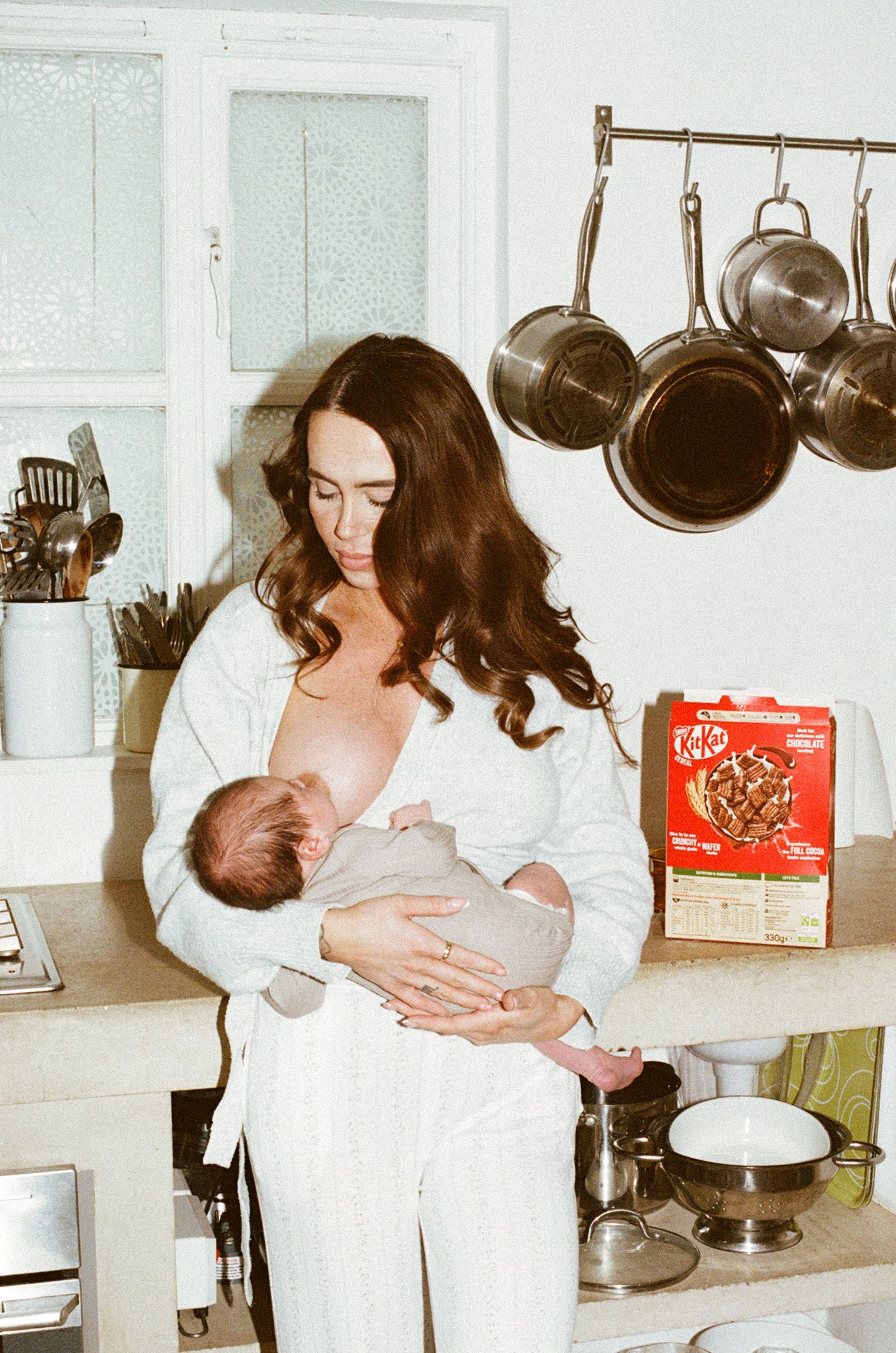 A mother breastfeeding her newborn baby in a cozy kitchen with hanging pots and a KitKat box nearby.