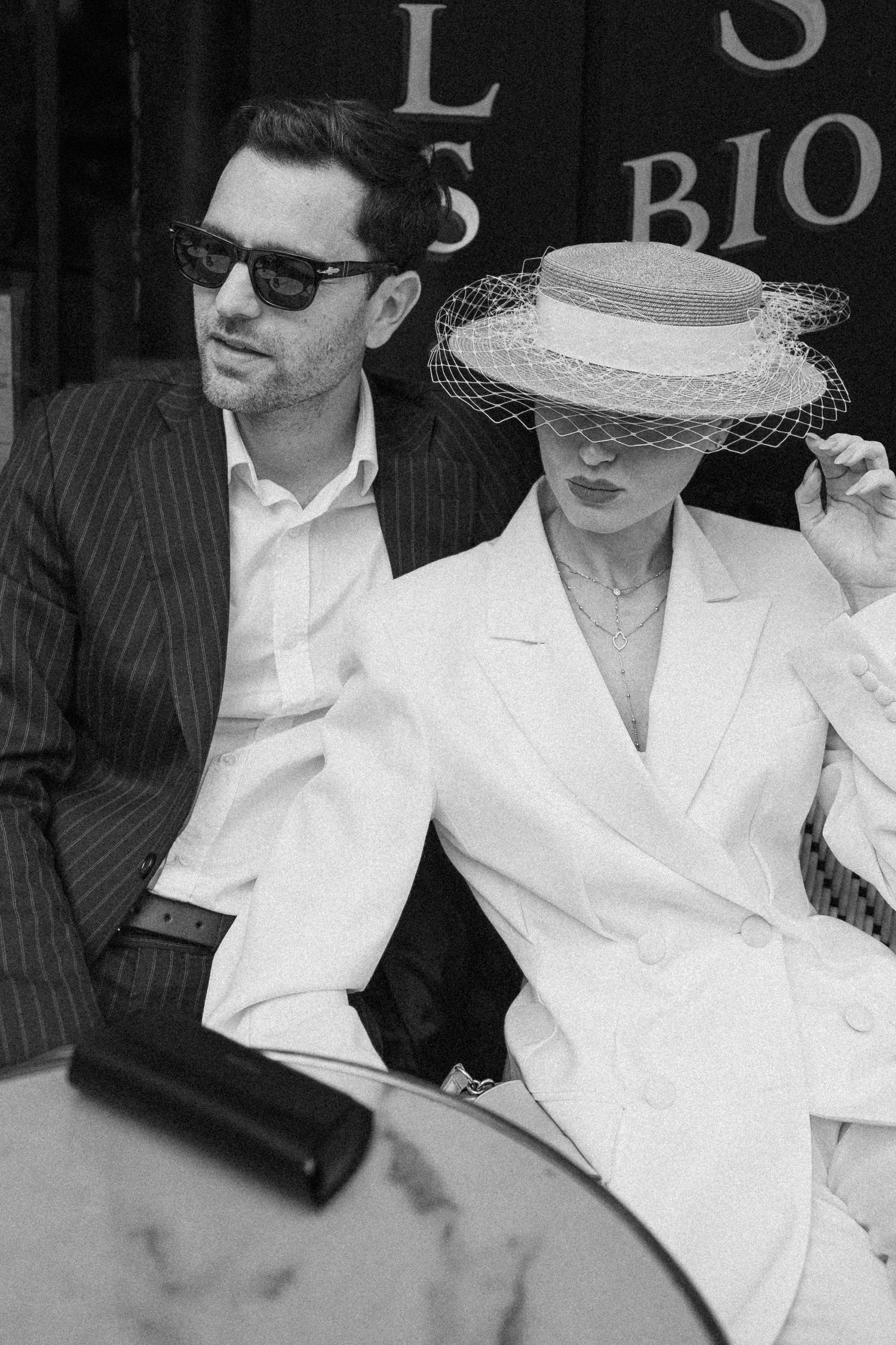 Black and white photo of an elegant couple, man in pinstripe suit with sunglasses, woman in white blazer and veiled boater hat.