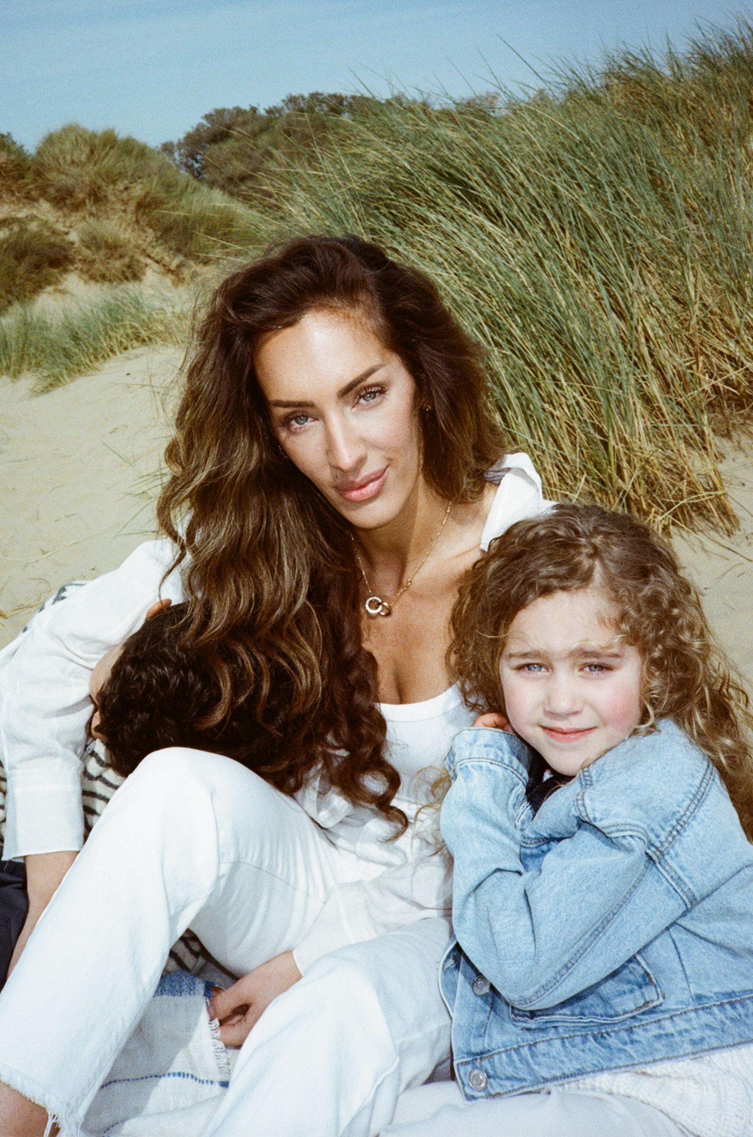 Woman and young girl sitting together on sandy beach dunes, both wearing white and denim outfits, smiling at camera.