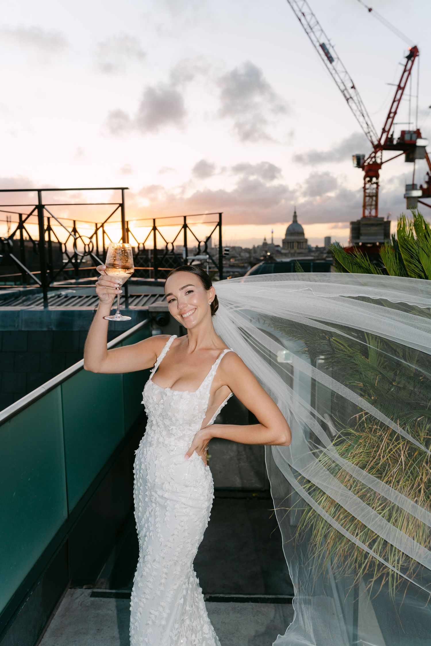 Bride dancing on rooftop terrace with city skyline and sunset in background.