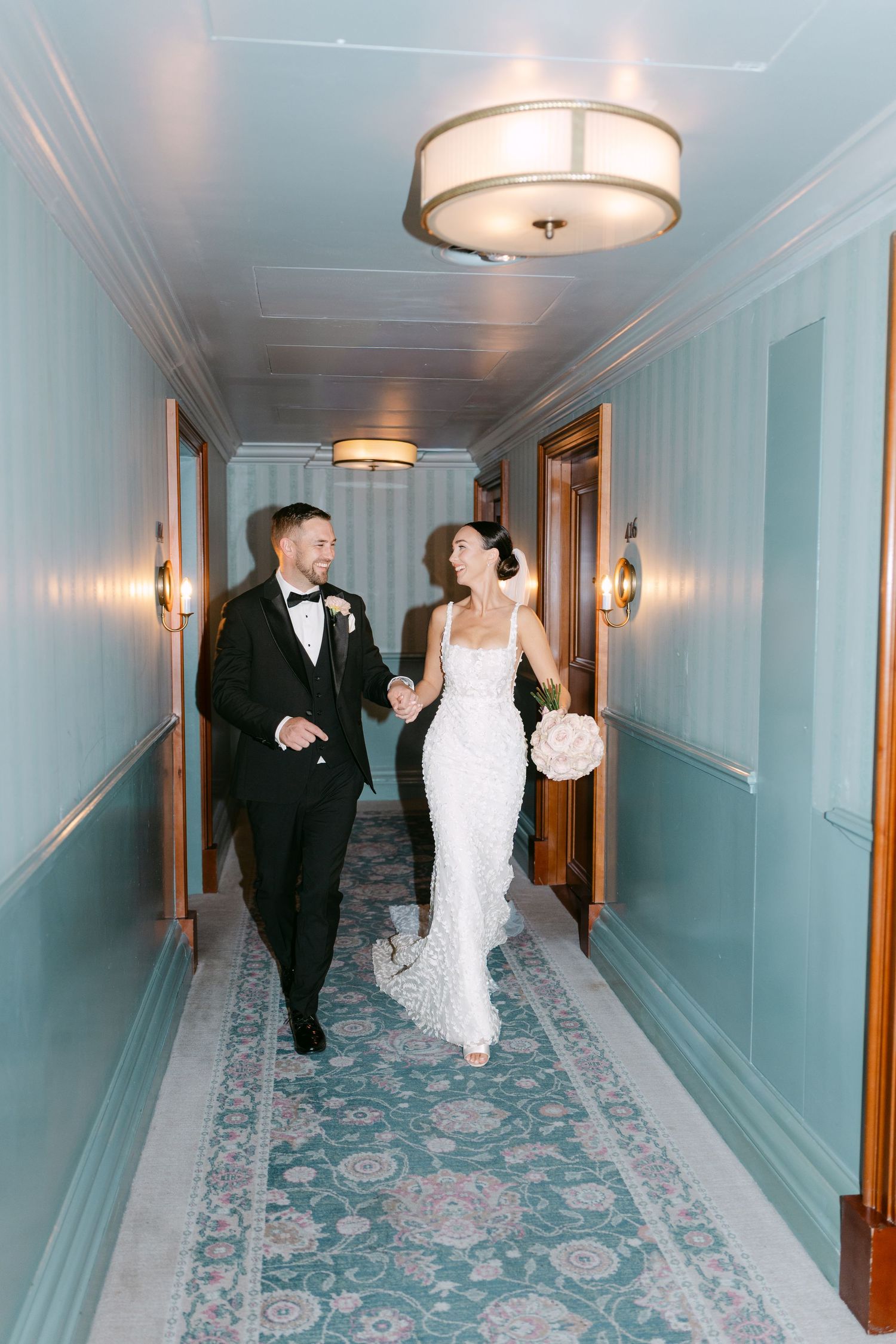 Couple walks playfully through mint-colored hotel corridor with patterned carpet.