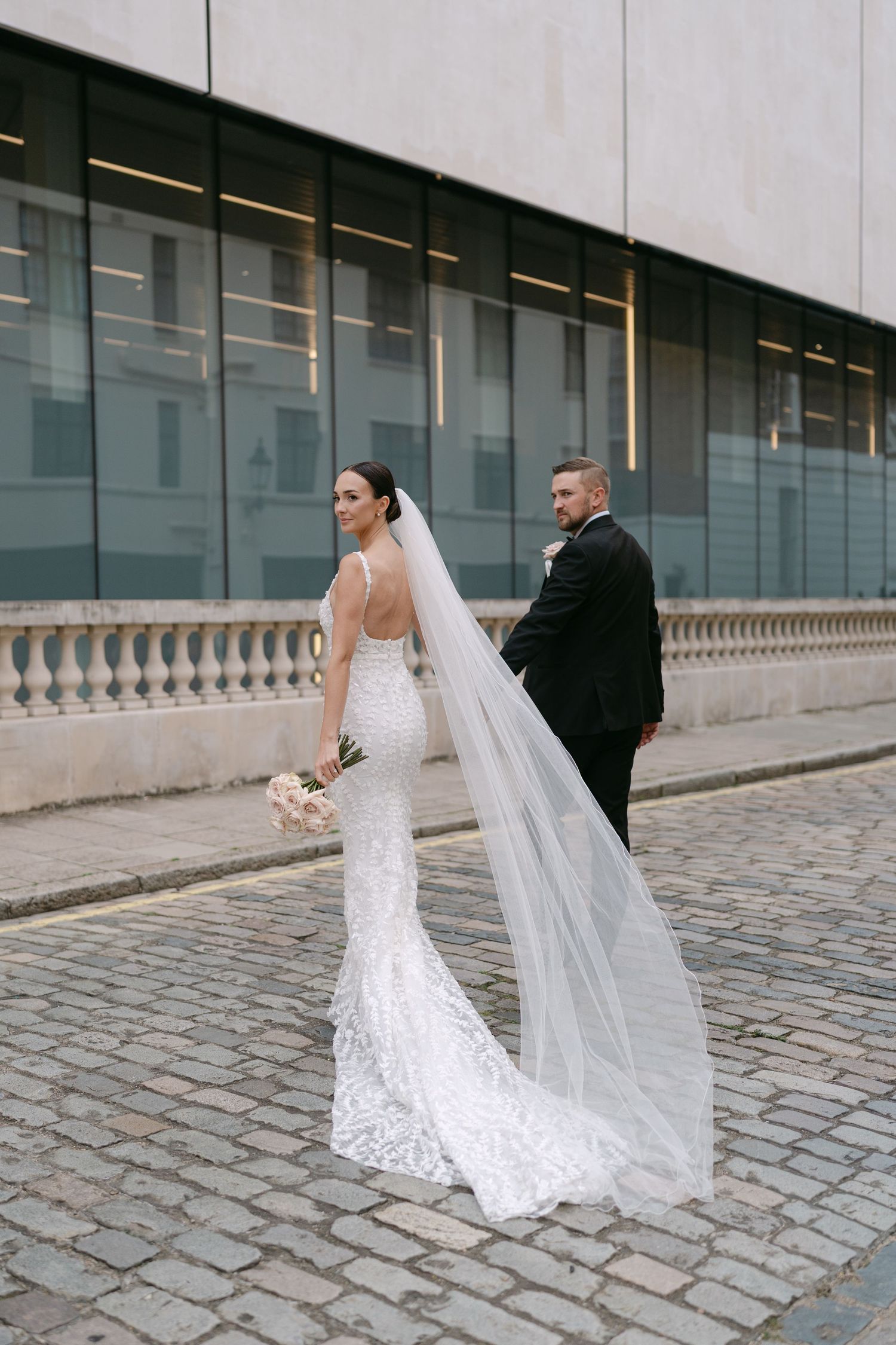 Wedding couple poses along modern glass building with flowing veil and cobblestone ground.