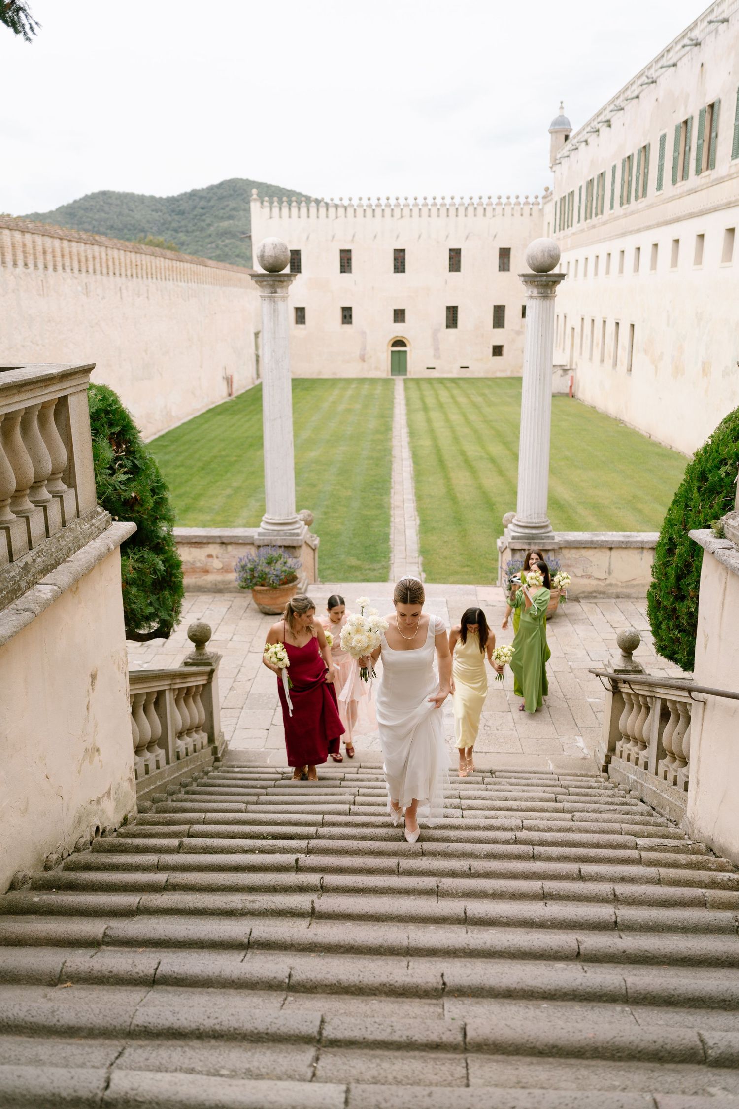 Wedding party descends grand stone staircase at historic castle with columns and green lawn.