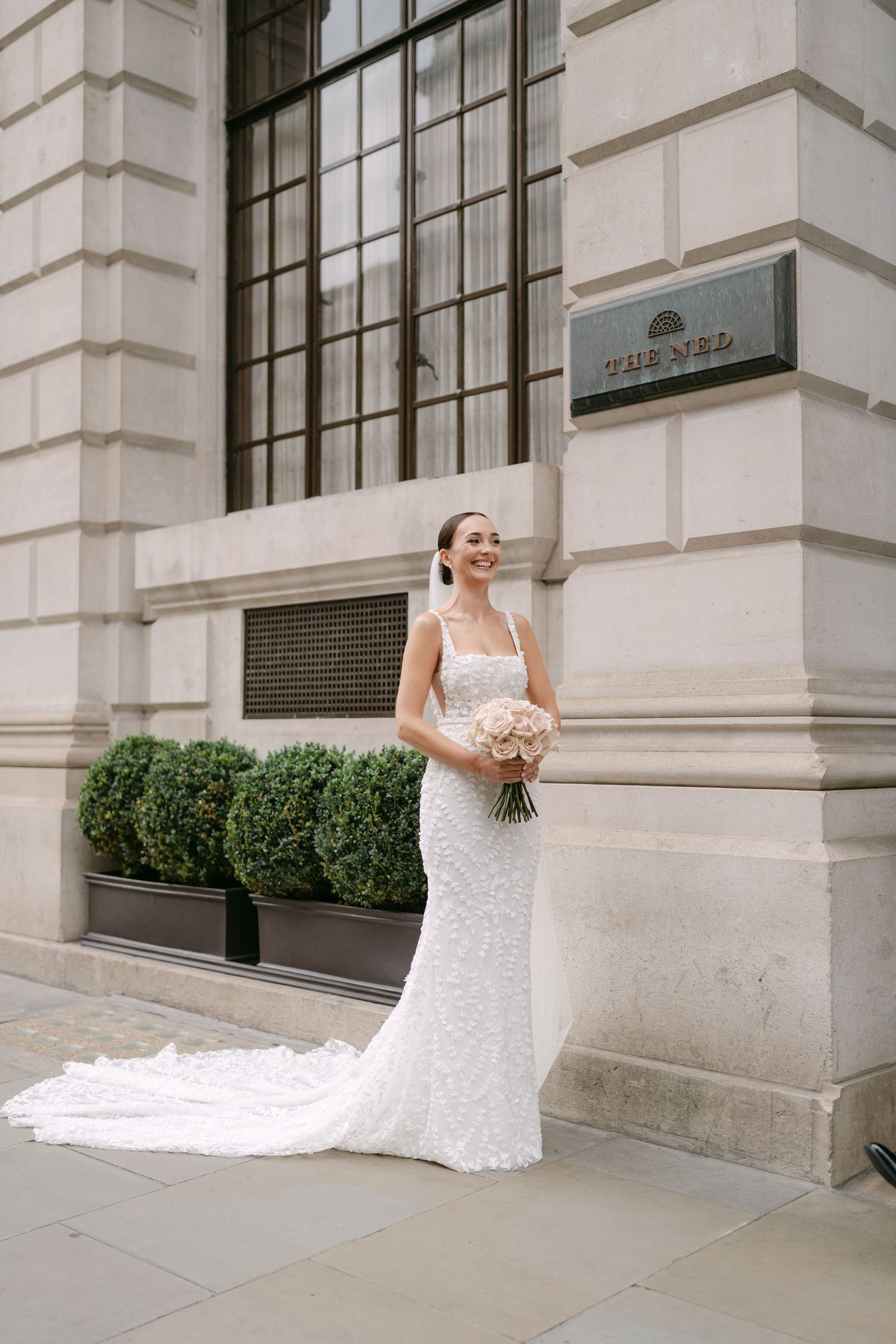 A stunning white lace wedding gown with a long train photographed against a classic stone building exterior.