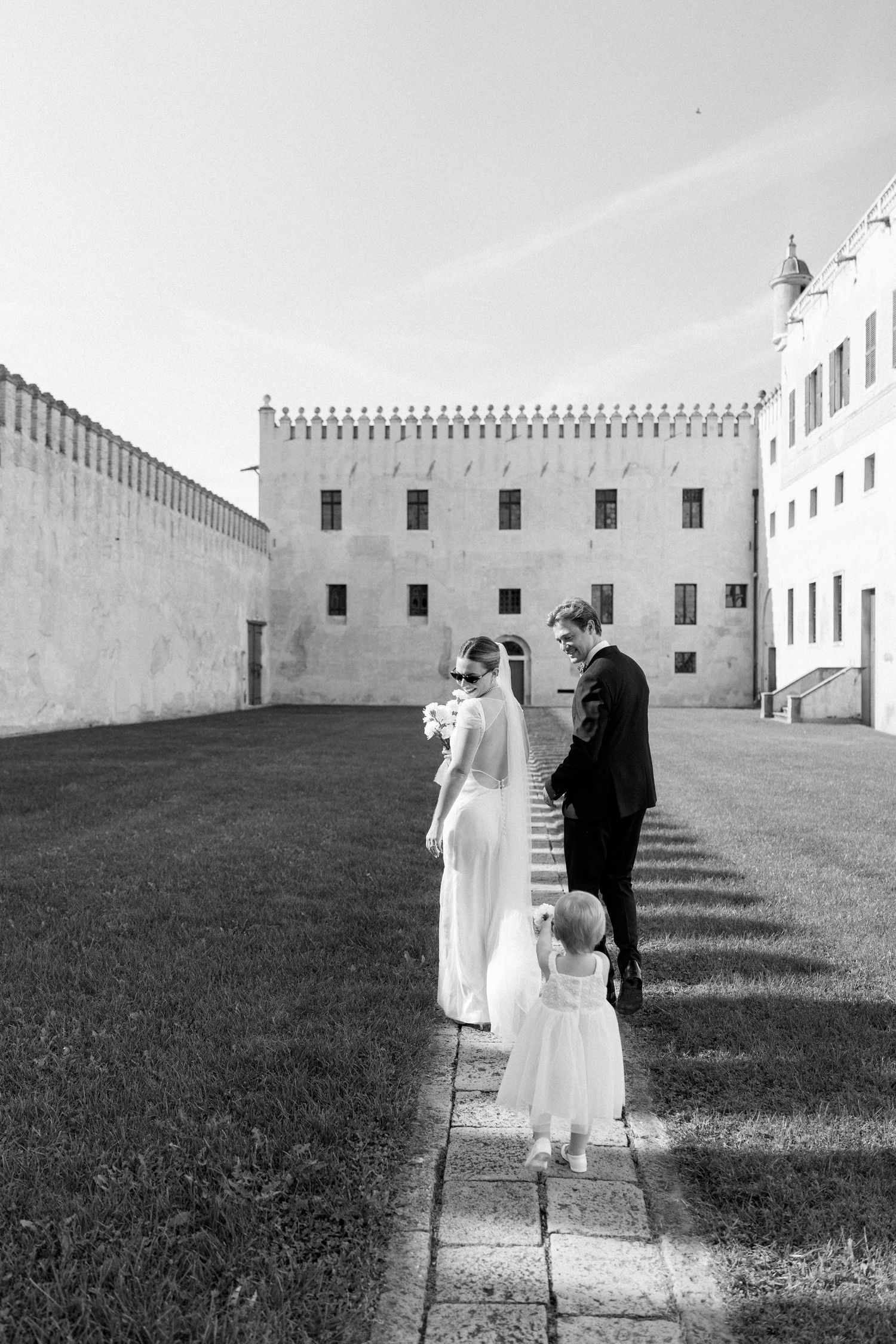 Wedding couple and child stand on stone path against white castle wall in black and white photograph.