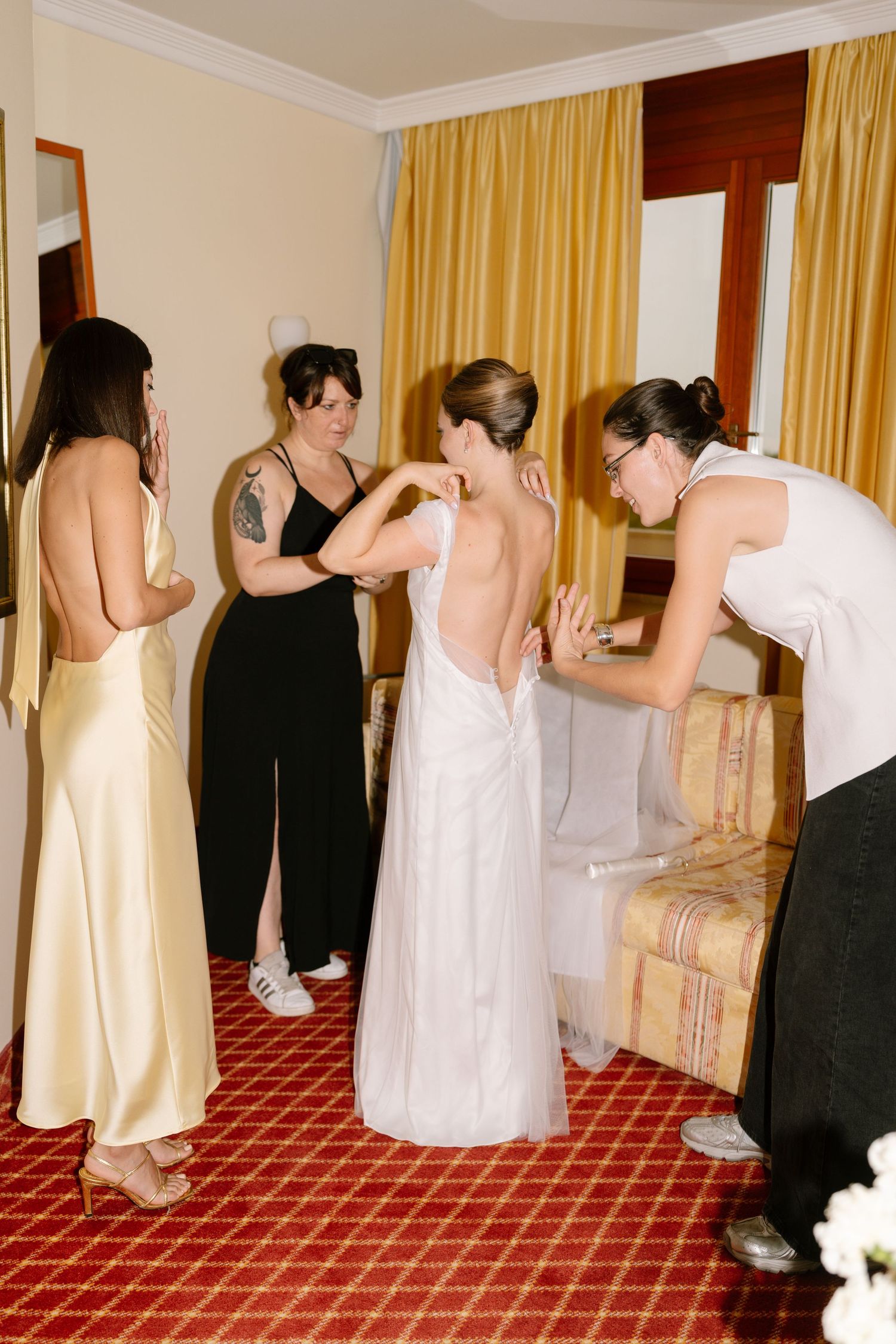 Wedding party members get ready in a hotel room with yellow curtains and red carpeting.